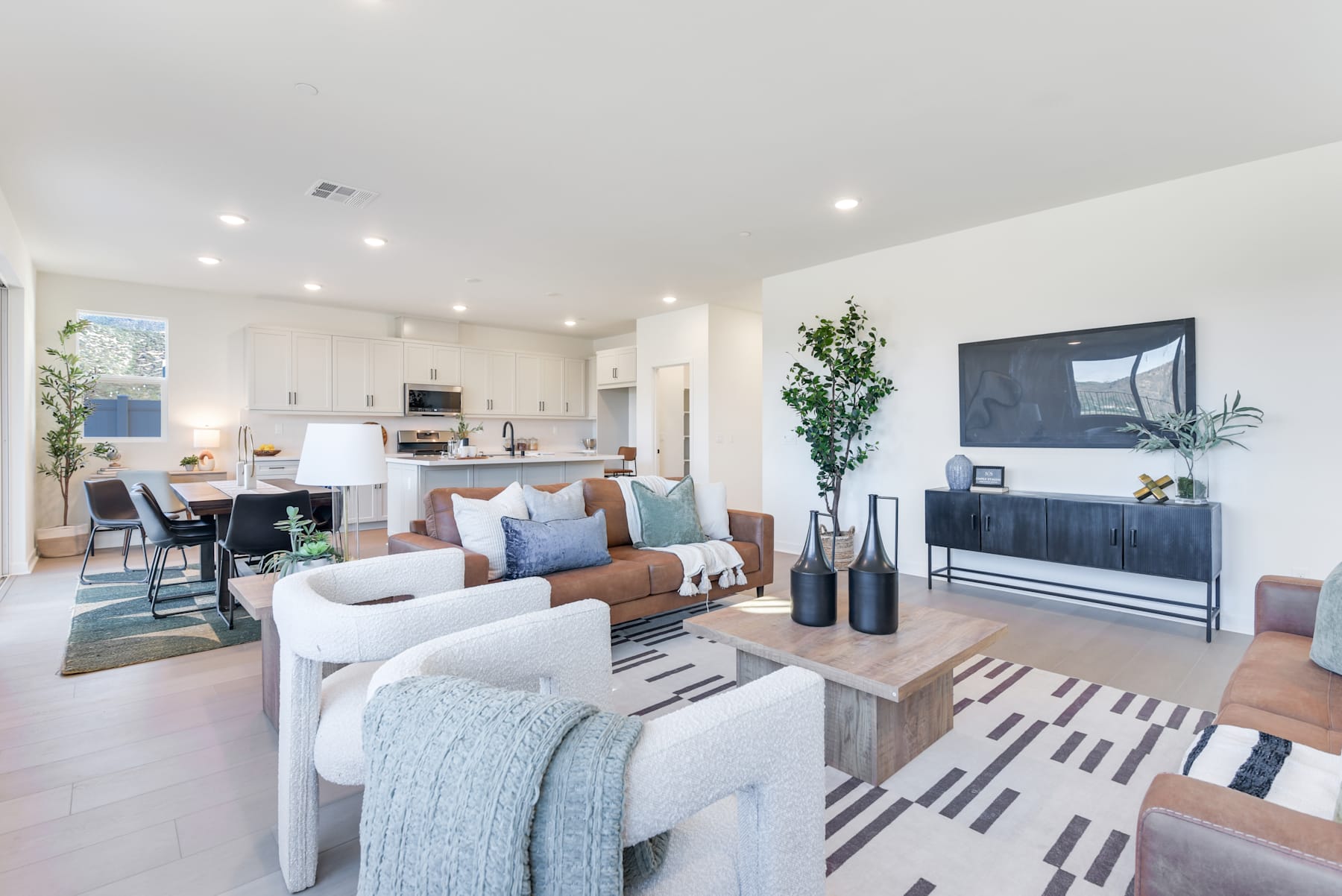 A modern and cozy living room with a white sofa, a wooden coffee table, and a TV stand with decorative items, surrounded by a bright and airy kitchen area.