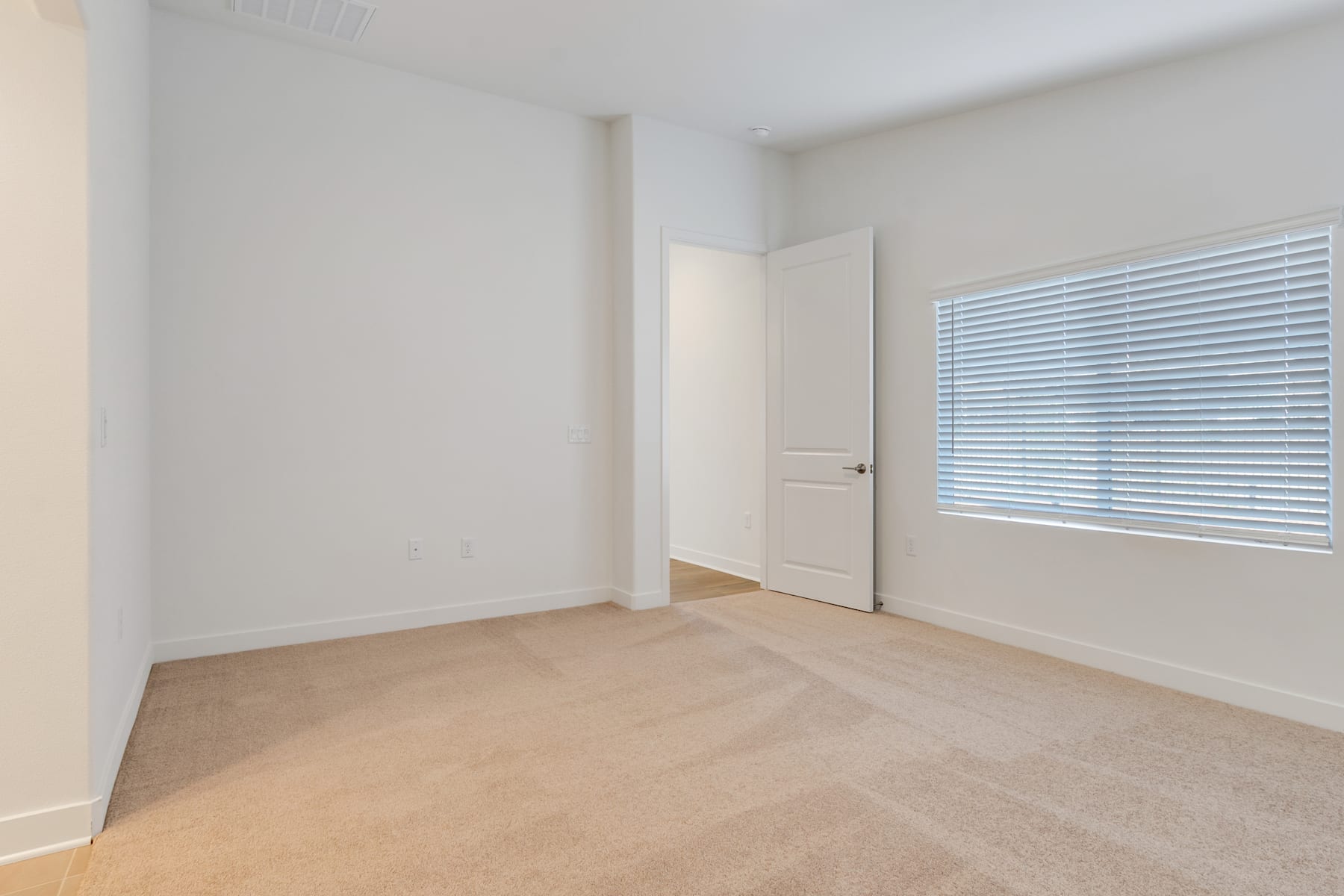 A simple, minimalist bedroom with white walls, a window with blinds, and a carpeted floor.