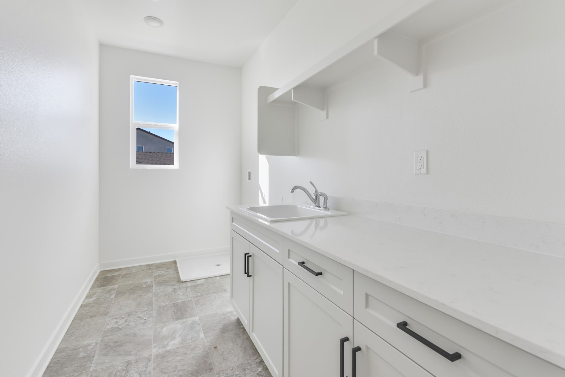 A modern, minimalist kitchen with white walls, gray cabinets, and a small window providing natural light.