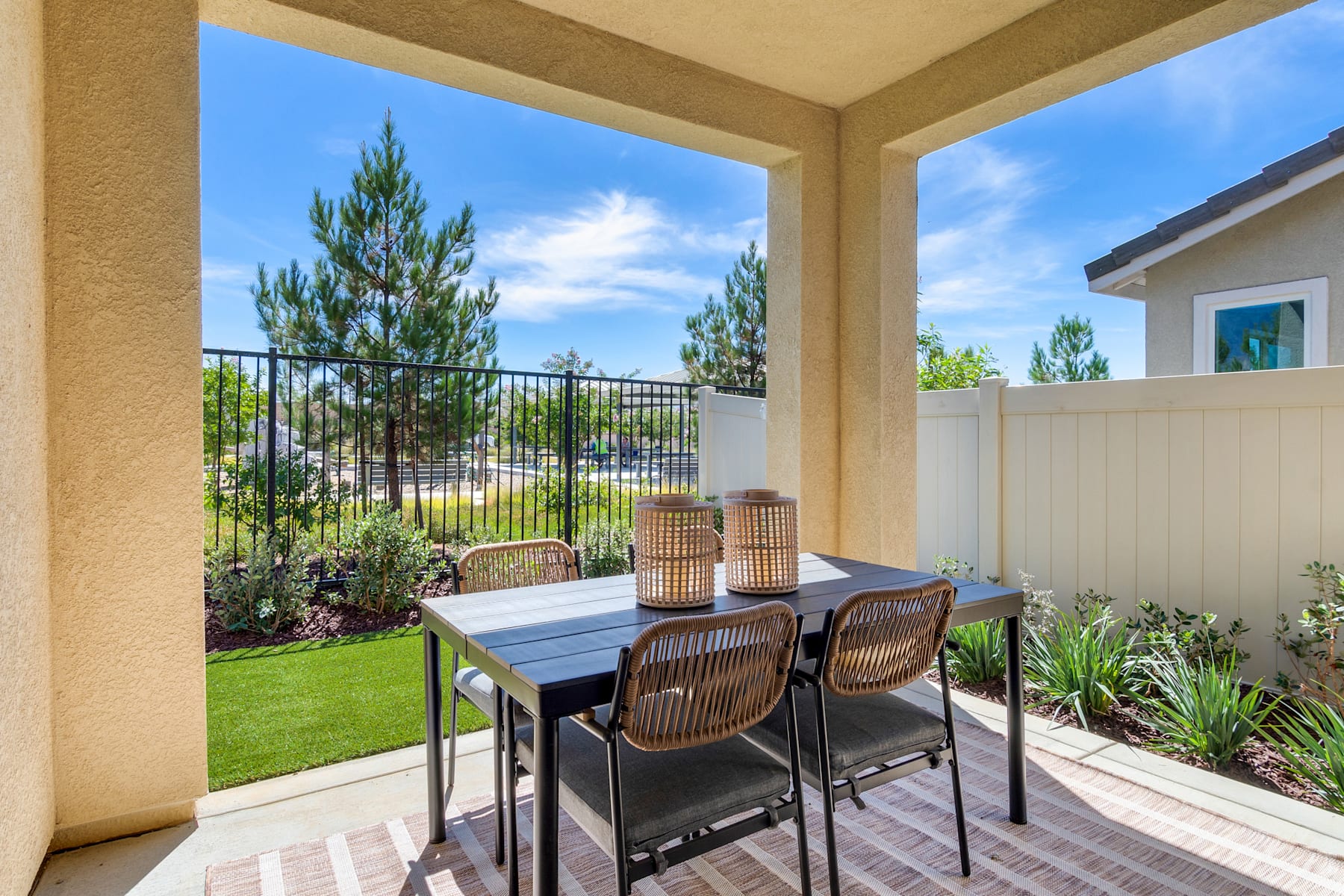 A cozy outdoor patio with a dining table and chairs, surrounded by lush greenery and a well-manicured lawn, with a clear blue sky visible in the background.