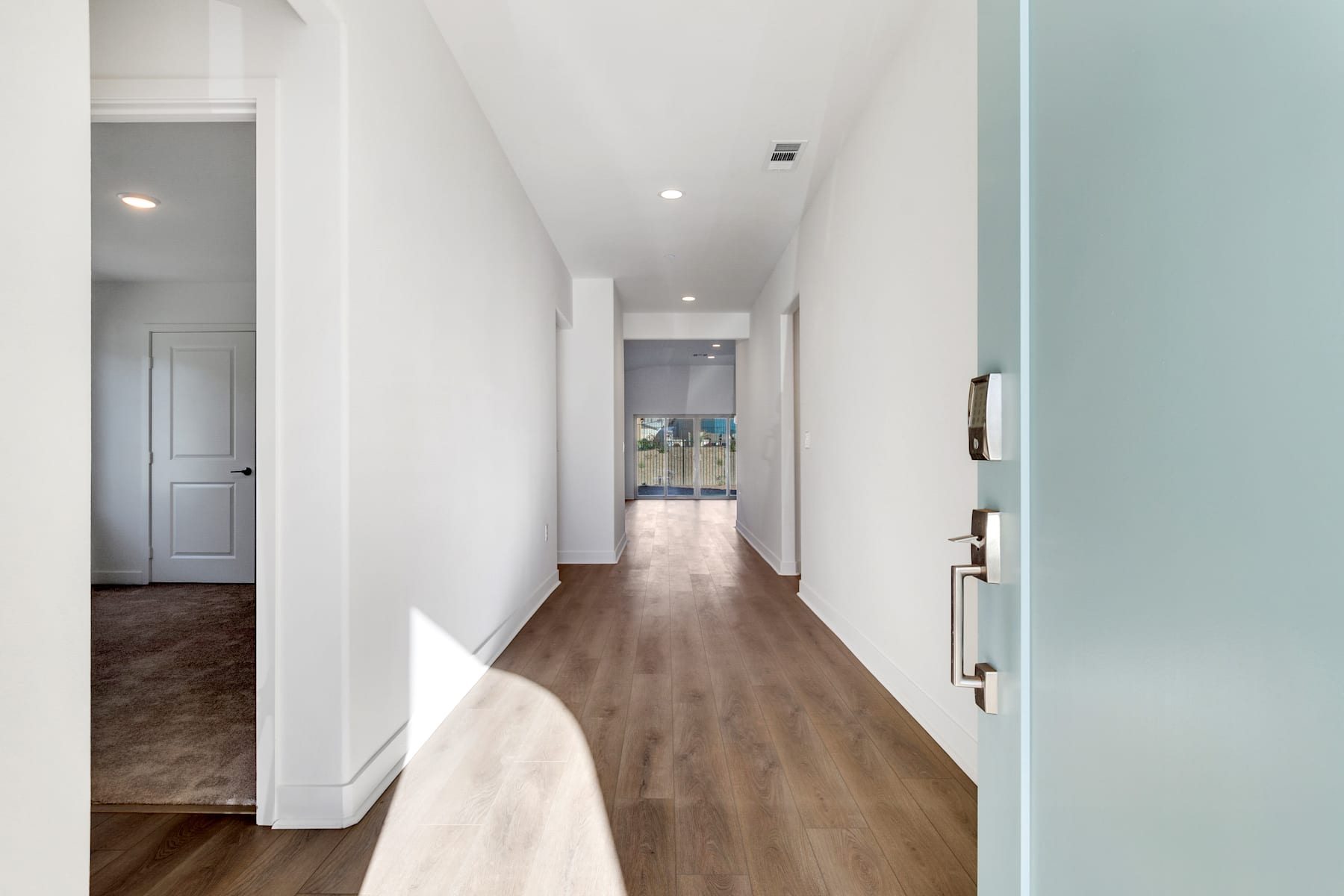 A long, well-lit hallway with white walls, a wooden floor, and a partially visible doorway leading to another room.
