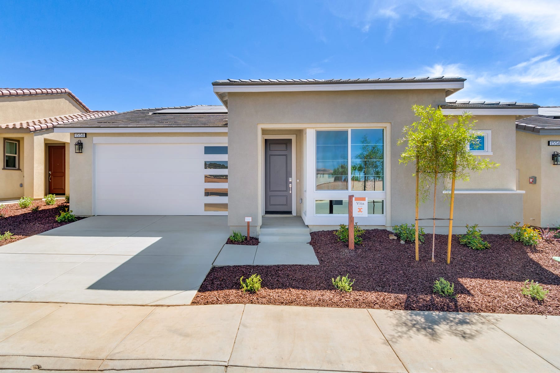 A modern, single-story house with a well-landscaped front yard, featuring a paved driveway, a small tree, and various shrubs and plants.