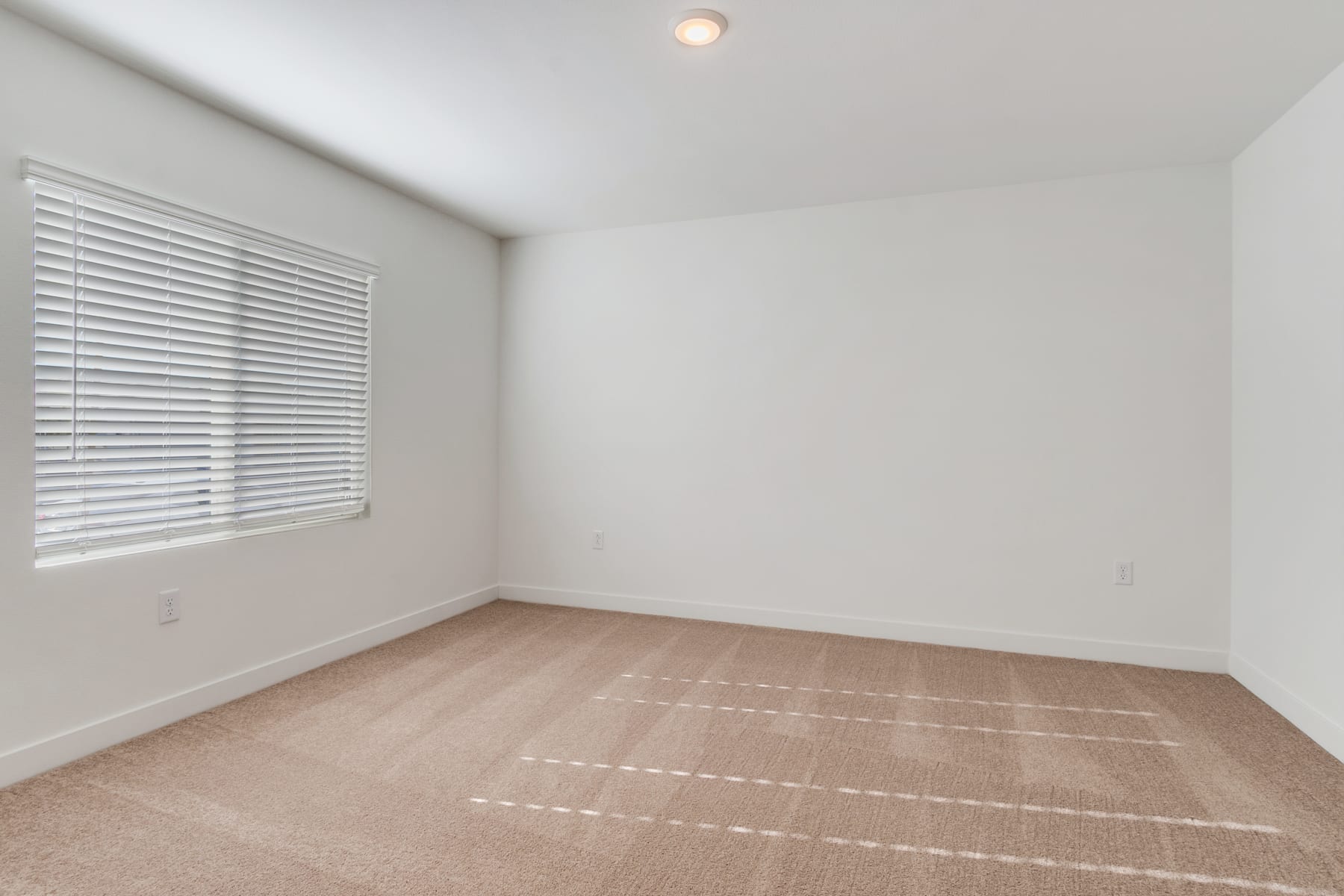 A bright, minimalist room with white walls, a window with horizontal blinds, and a hardwood floor.