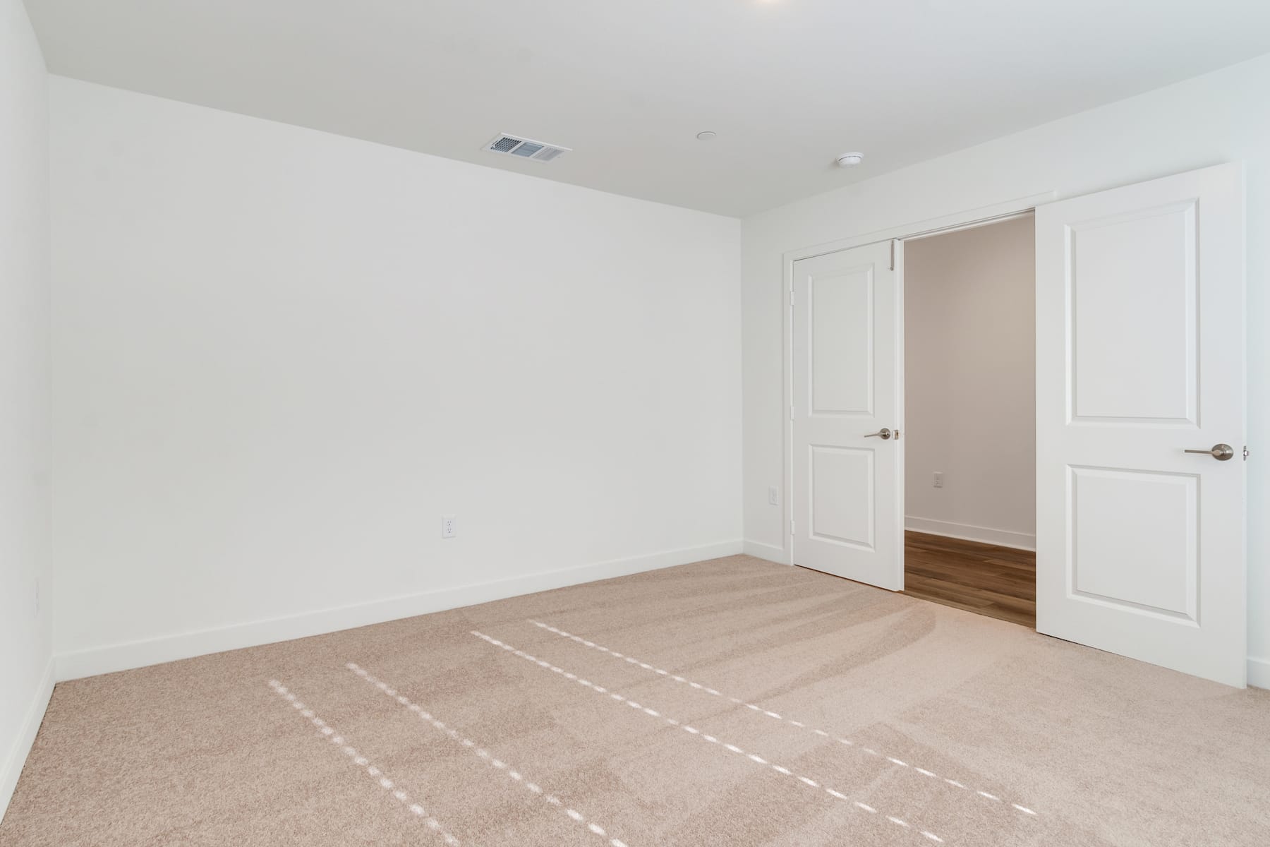 A simple, minimalist bedroom with white walls, a beige carpeted floor, and two closed doors leading to other rooms.