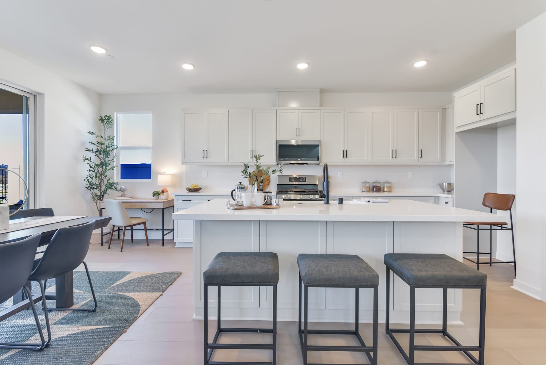 A modern, open-concept kitchen with white cabinets, a large island, and bar stools, set against a backdrop of large windows overlooking a scenic view.