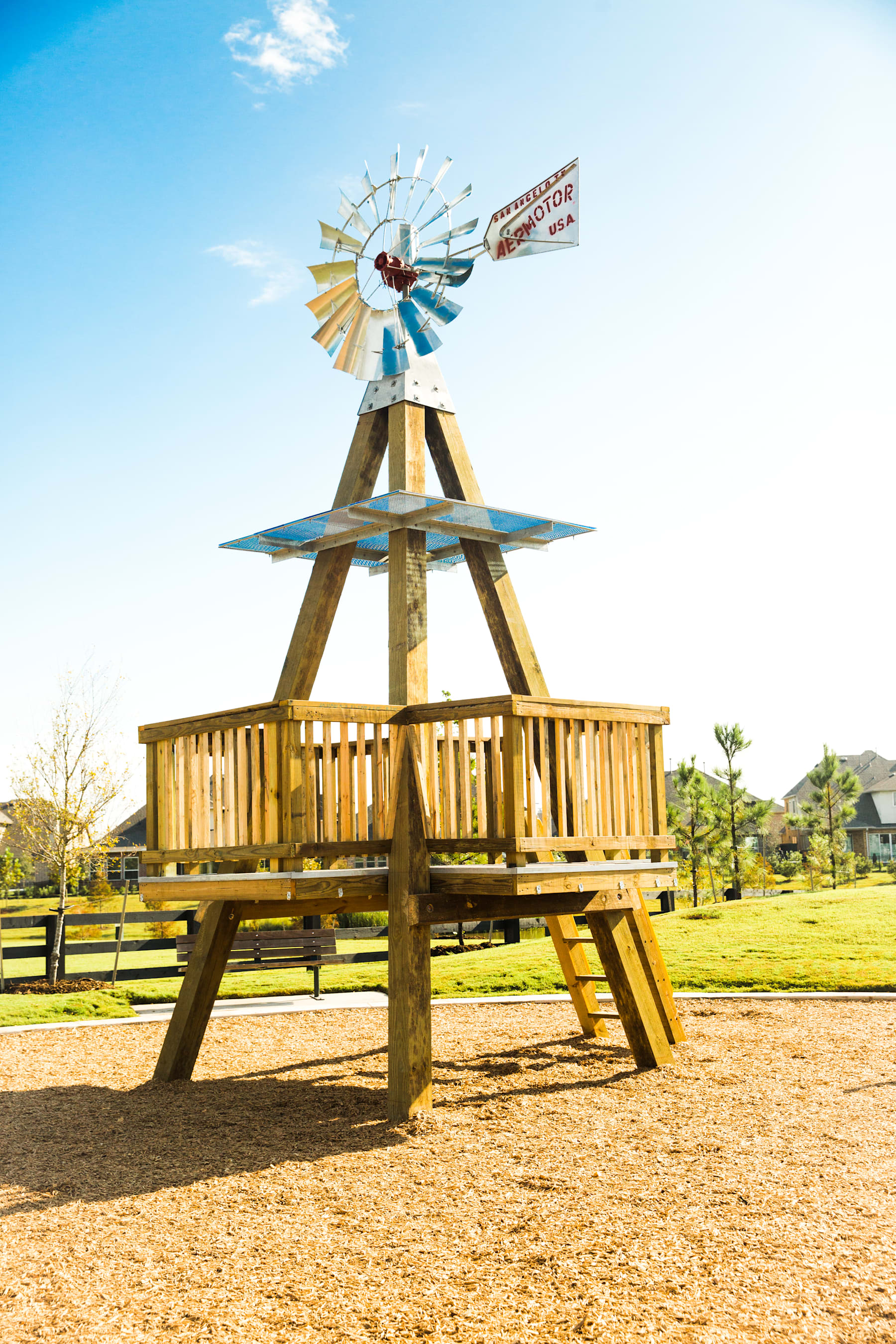 A wooden playground structure with a colorful windmill on top stands in a grassy field against a blue sky with scattered clouds.