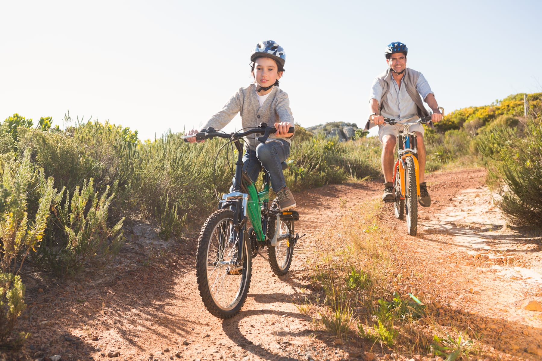 Two people riding bicycles on a dirt trail surrounded by lush vegetation and a clear sky.