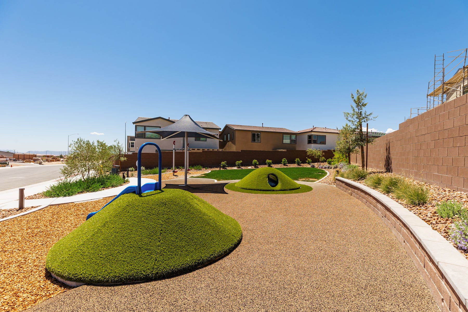 A residential neighborhood with a well-designed landscaped area featuring sculpted grass mounds, a paved walkway, and a playground in the foreground, with houses and a clear blue sky in the background.