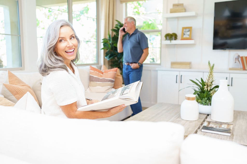 A smiling woman sits in a bright, modern living room, while a man stands in the background.