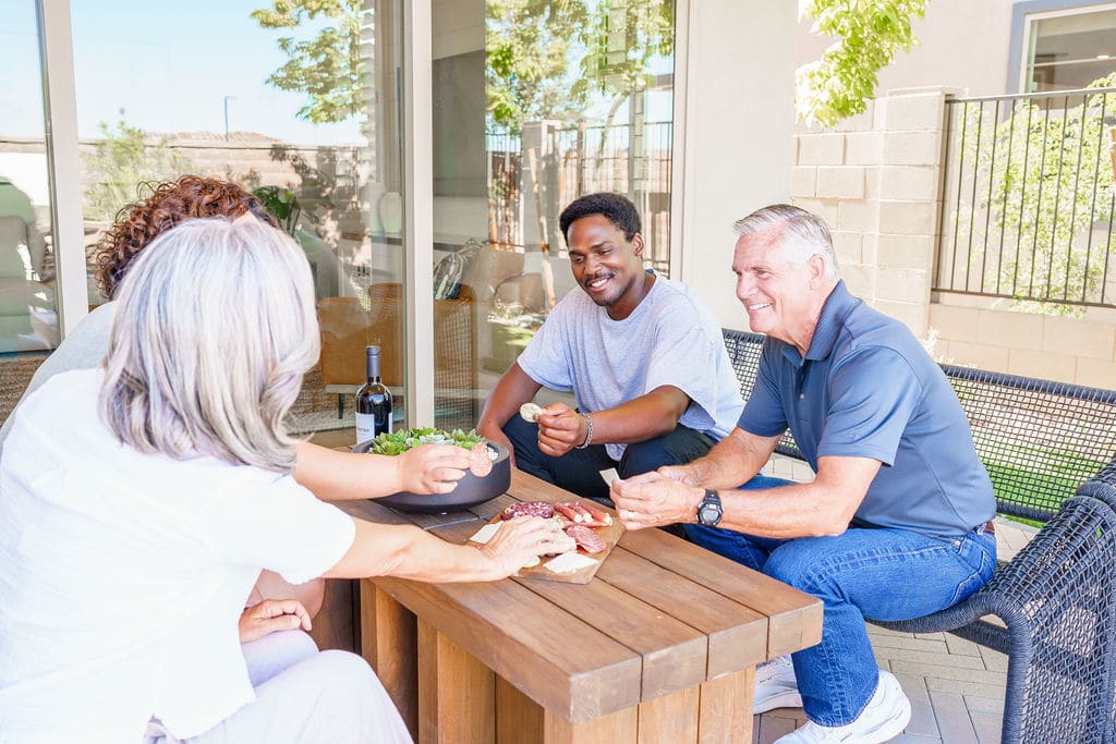 A group of people, including a woman in a white dress, are gathered around a table on an outdoor patio, engaged in conversation and enjoying a glass of wine.