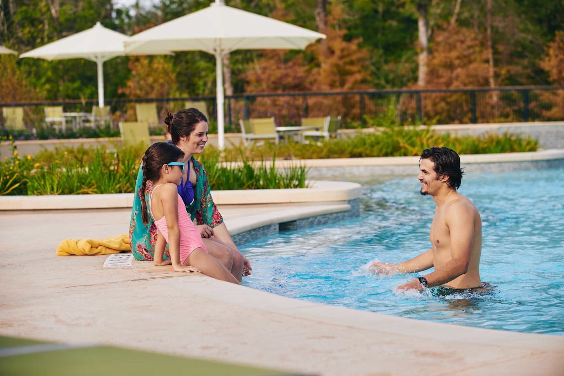 The image shows a group of people relaxing by a swimming pool, with a gazebo and lush greenery in the background.