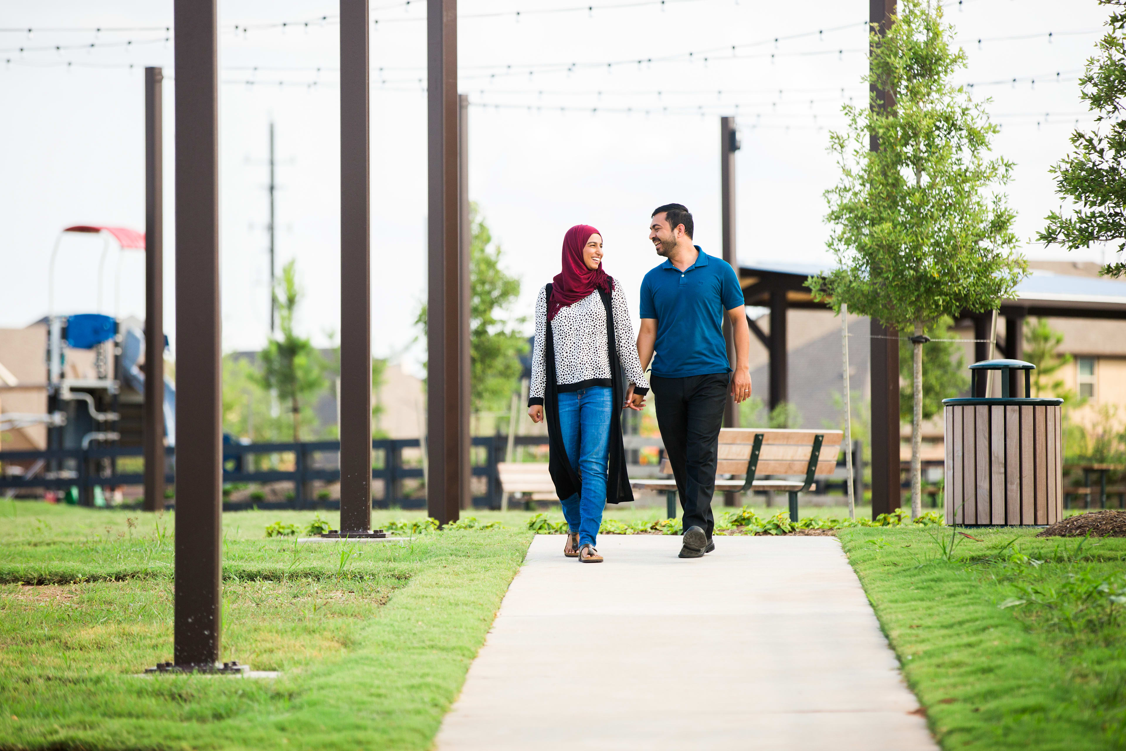 Two people, a man and a woman, are walking together on a paved path surrounded by lush greenery and trees in an outdoor setting.