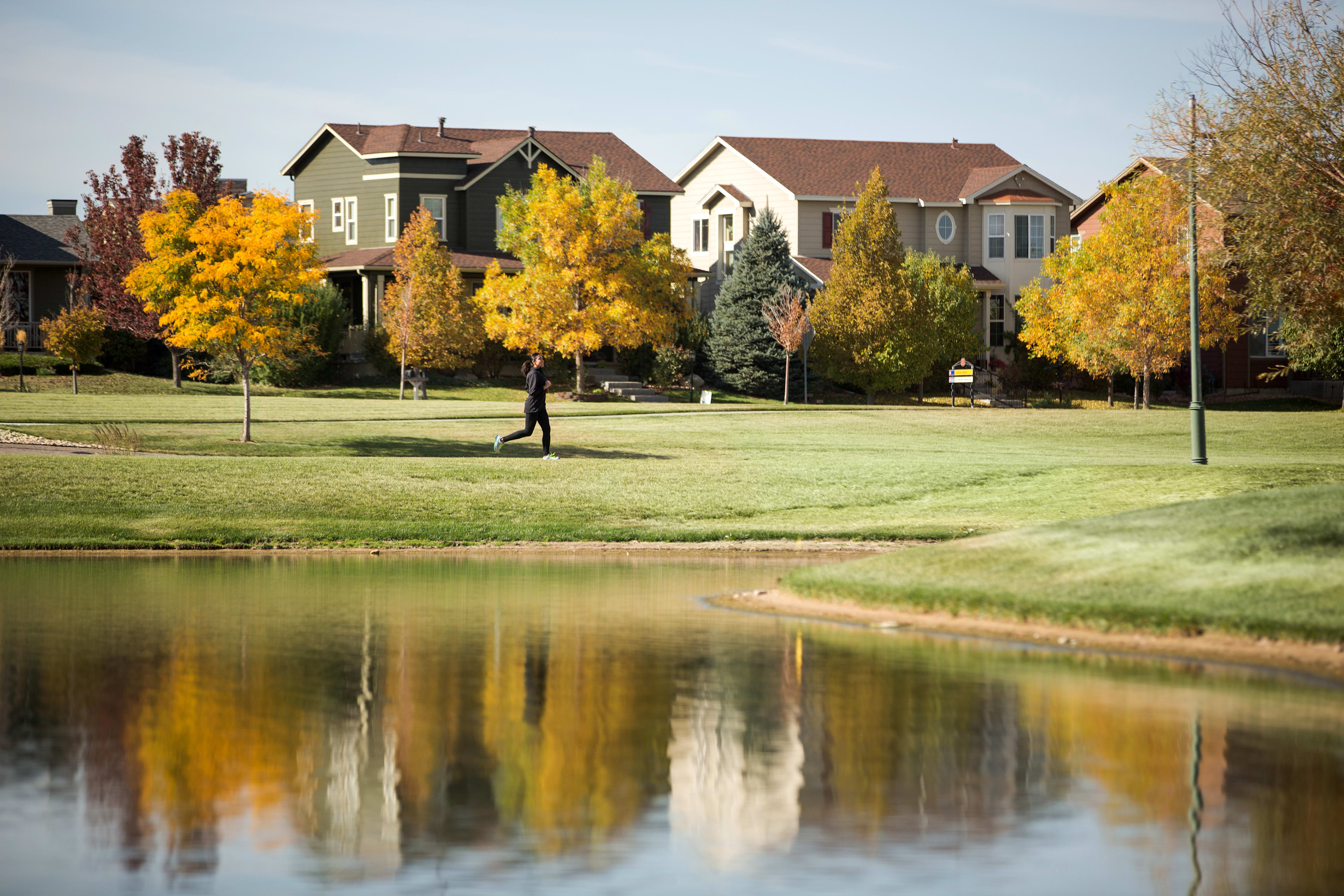 A peaceful suburban neighborhood with colorful autumn trees reflected in a tranquil pond, with a person walking in the foreground.