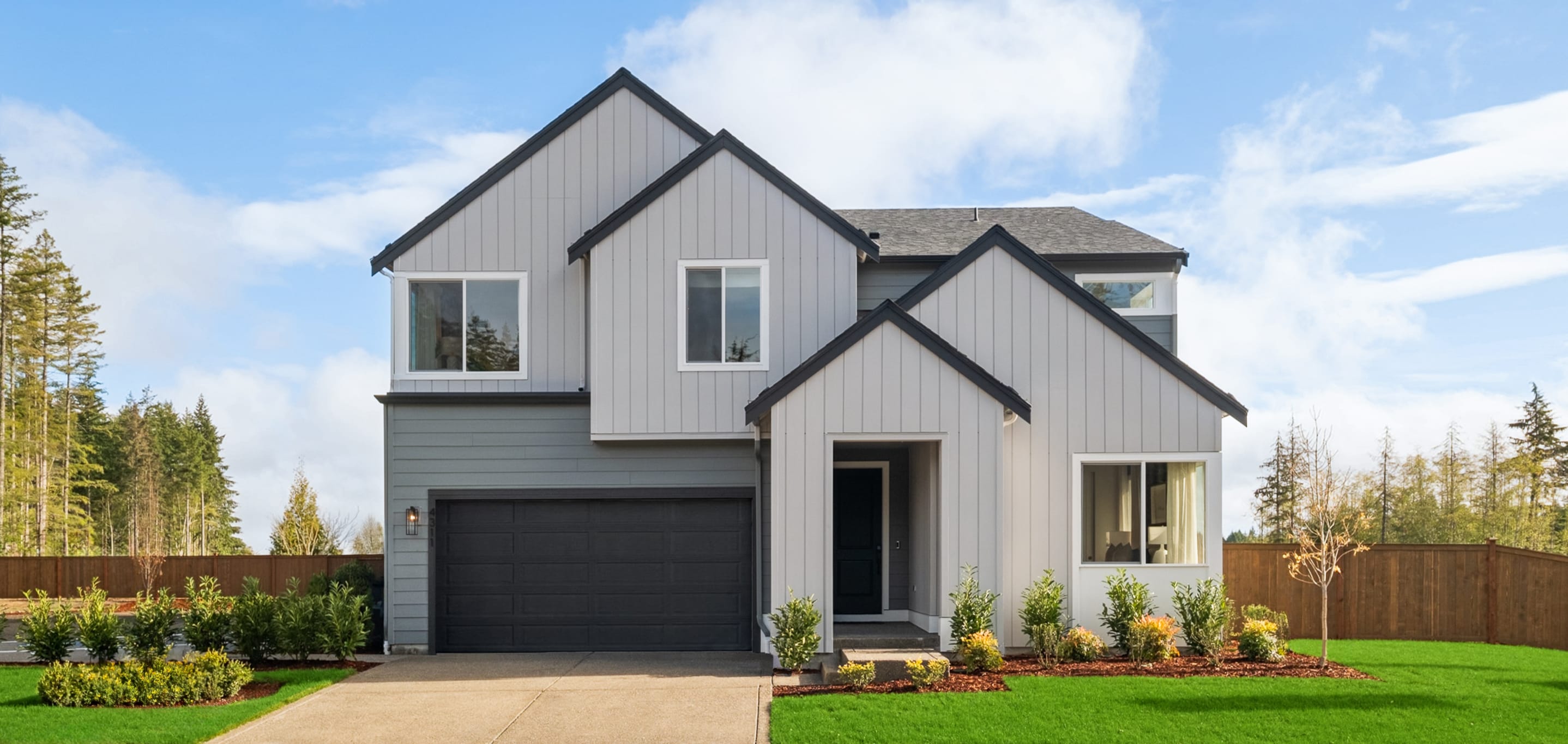 A modern two-story house with a gray exterior, a black garage door, and a well-manicured lawn surrounded by trees in the background.
