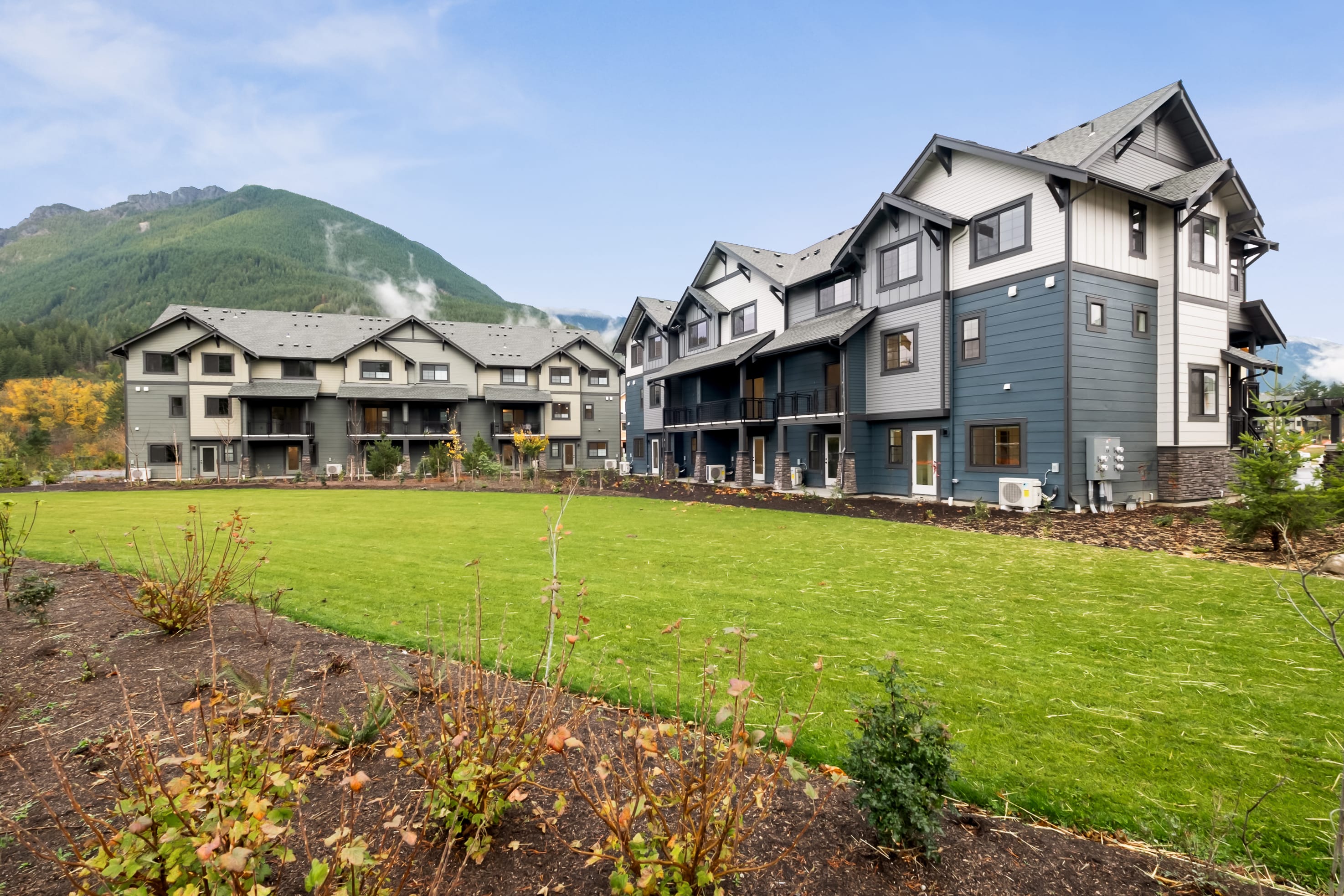 A group of modern, multi-story residential buildings with gabled roofs and gray siding, surrounded by a well-manicured grassy area and set against a backdrop of lush, green mountains.