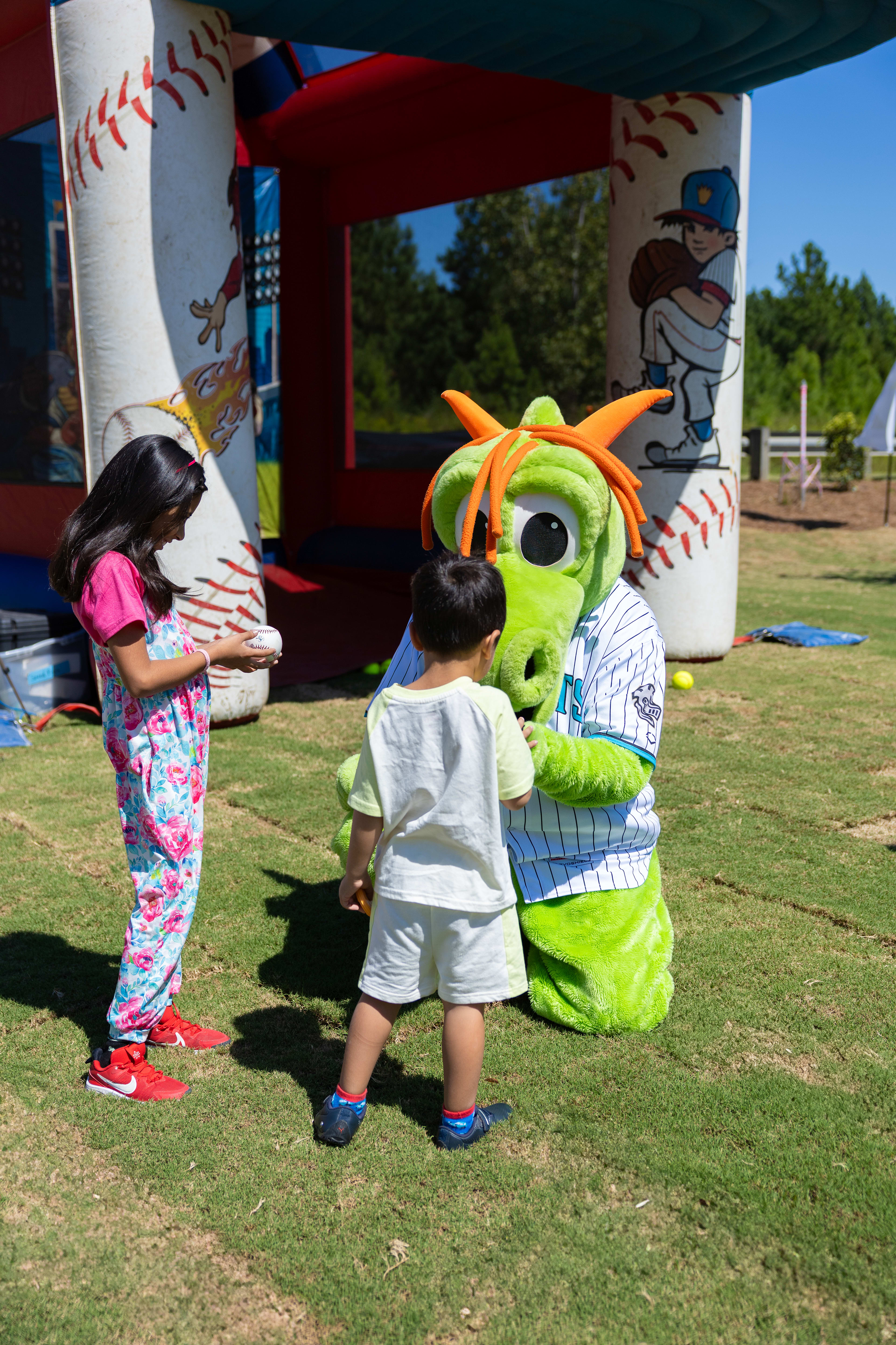 The image shows a large, inflatable green monster character interacting with two young children on a grassy field, with baseball-themed decorations and structures visible in the background.