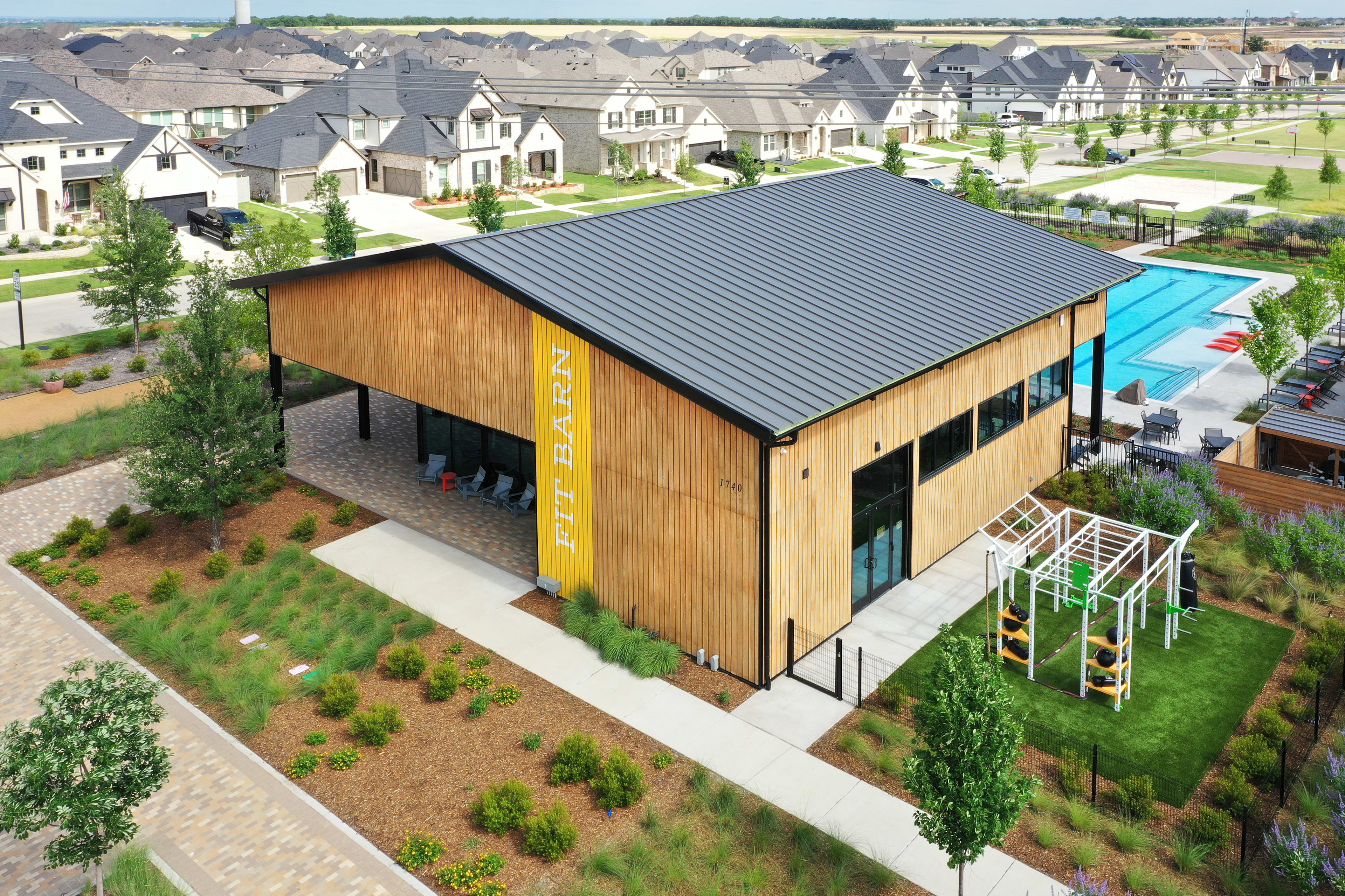 A modern, wooden-clad building with a black metal roof stands in the foreground, surrounded by a lush, grassy landscape. In the background, a residential neighborhood with rows of houses can be seen.