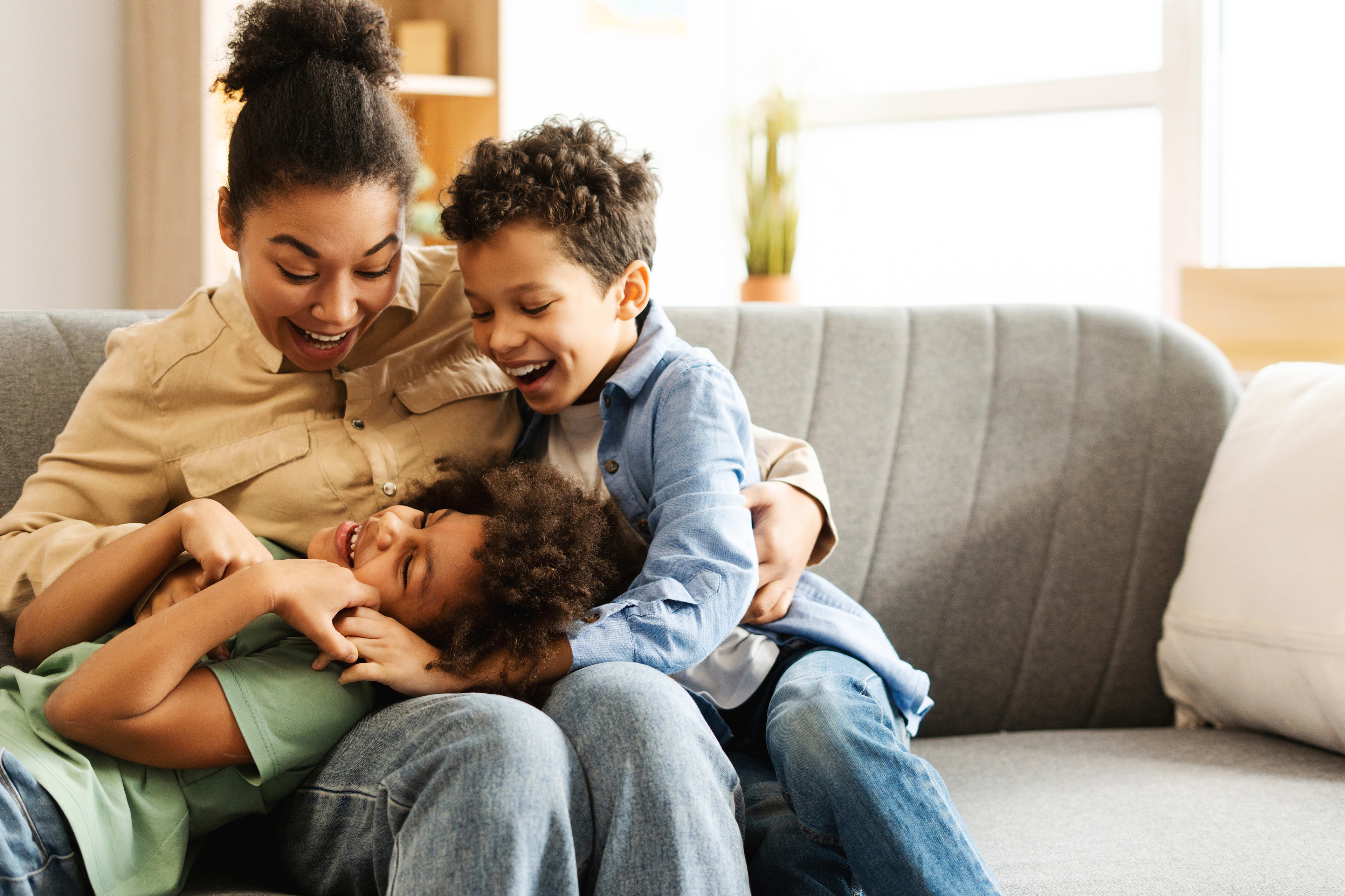 A joyful family moment captured, with a mother and two children cuddling and laughing together on a couch in a cozy, warm-toned living room setting.