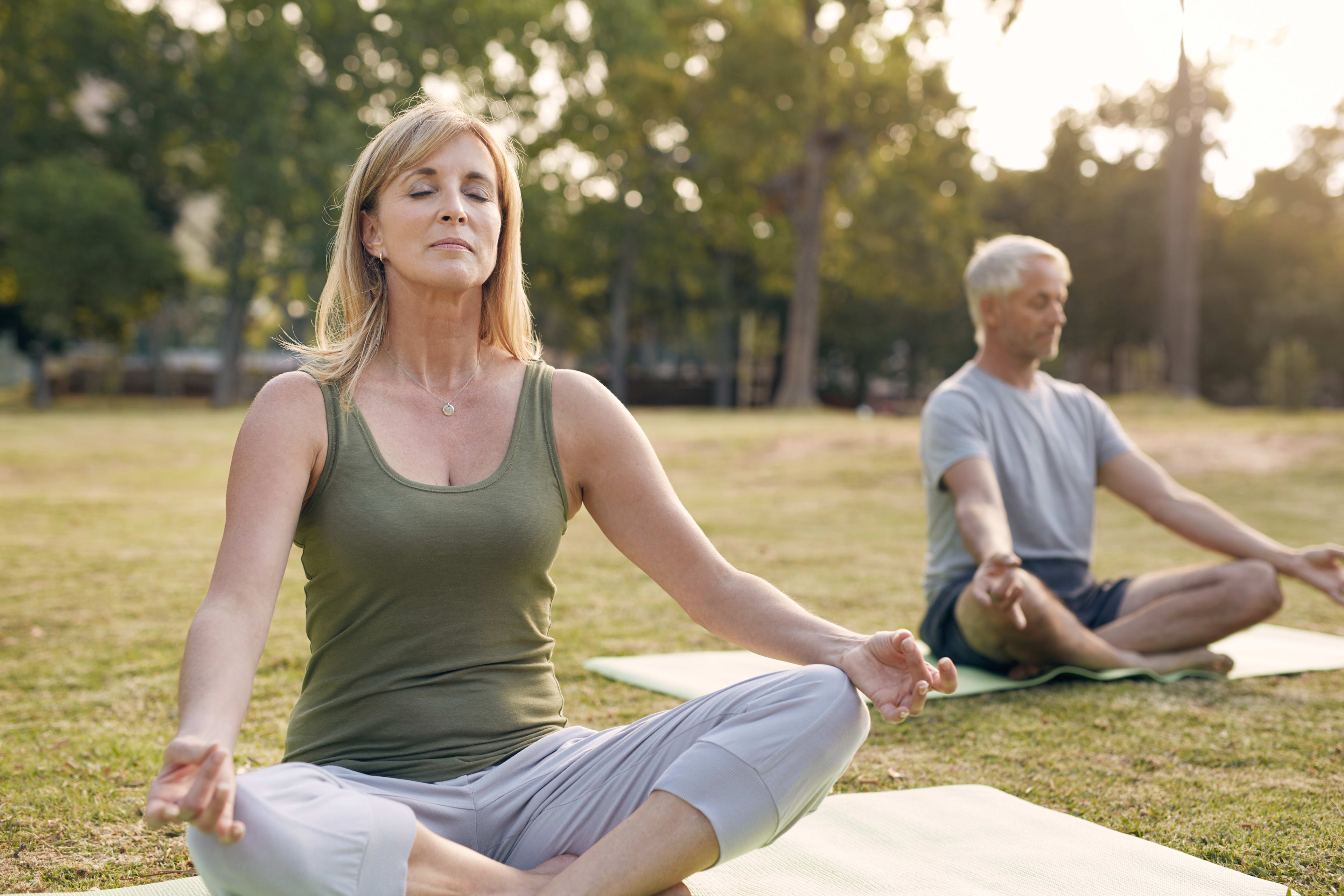 A woman in a green tank top and a man in a gray shirt are sitting in a meditative pose on yoga mats in a grassy park setting with trees in the background.