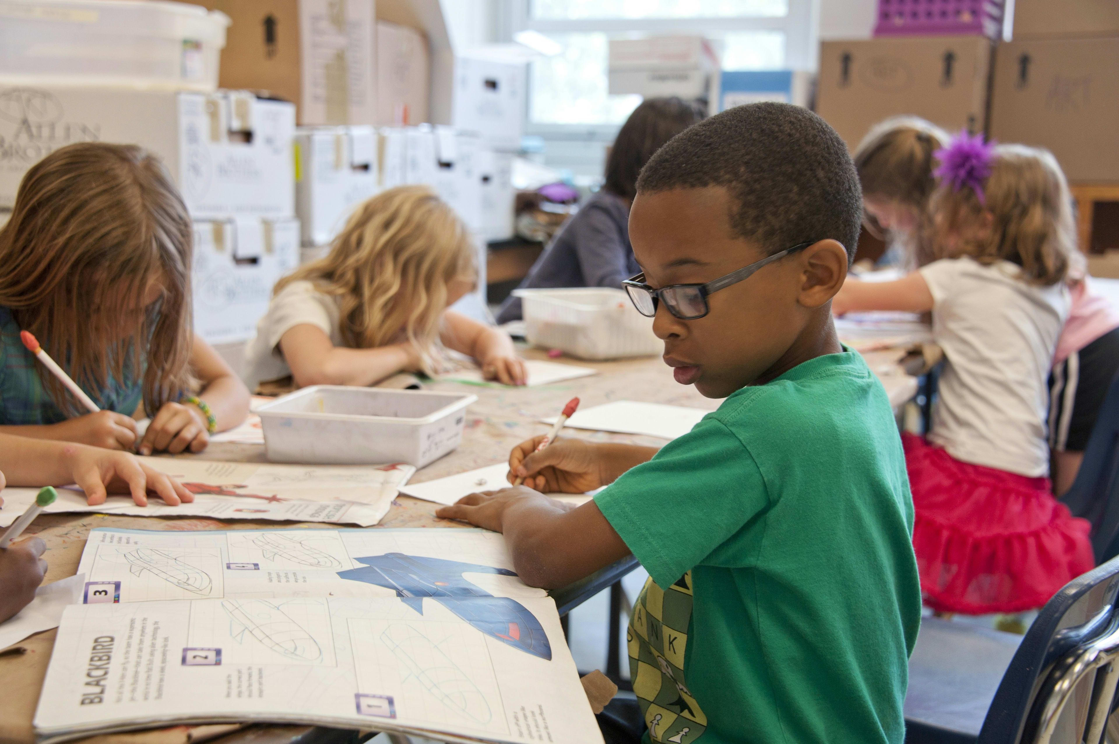 A group of students, both boys and girls, are engaged in various activities at their desks in a classroom setting, with posters and other educational materials visible in the background.