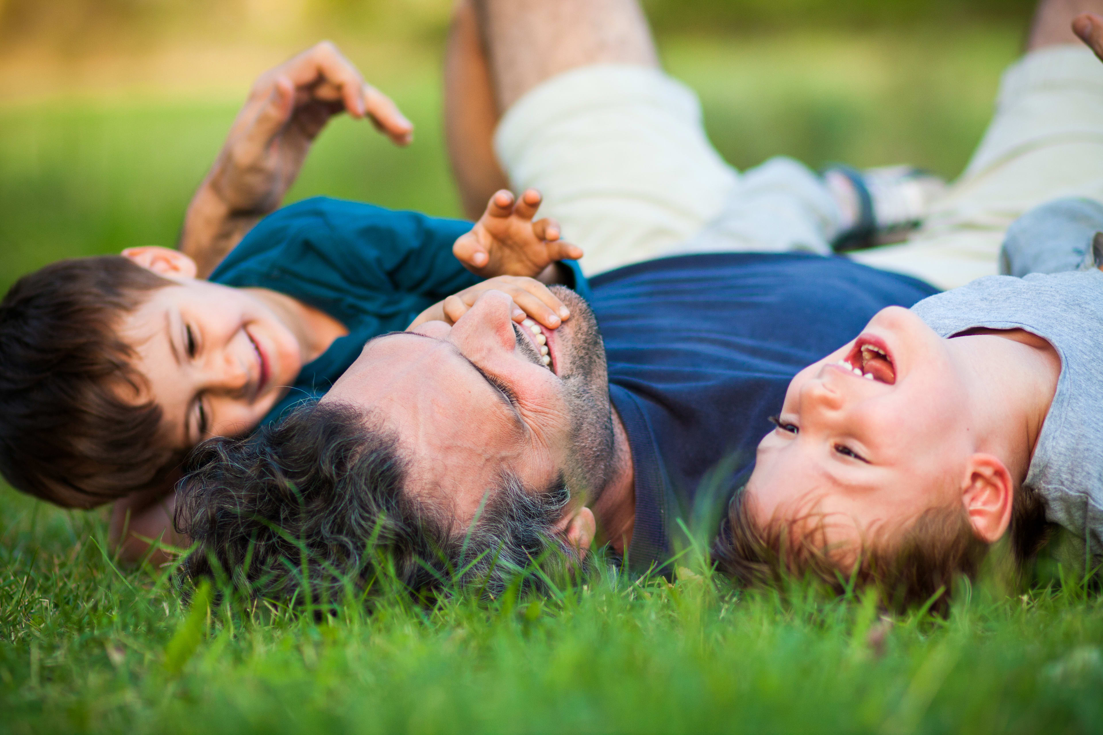 A family of three, a father and two children, lying on the grass in a lush, green outdoor setting.