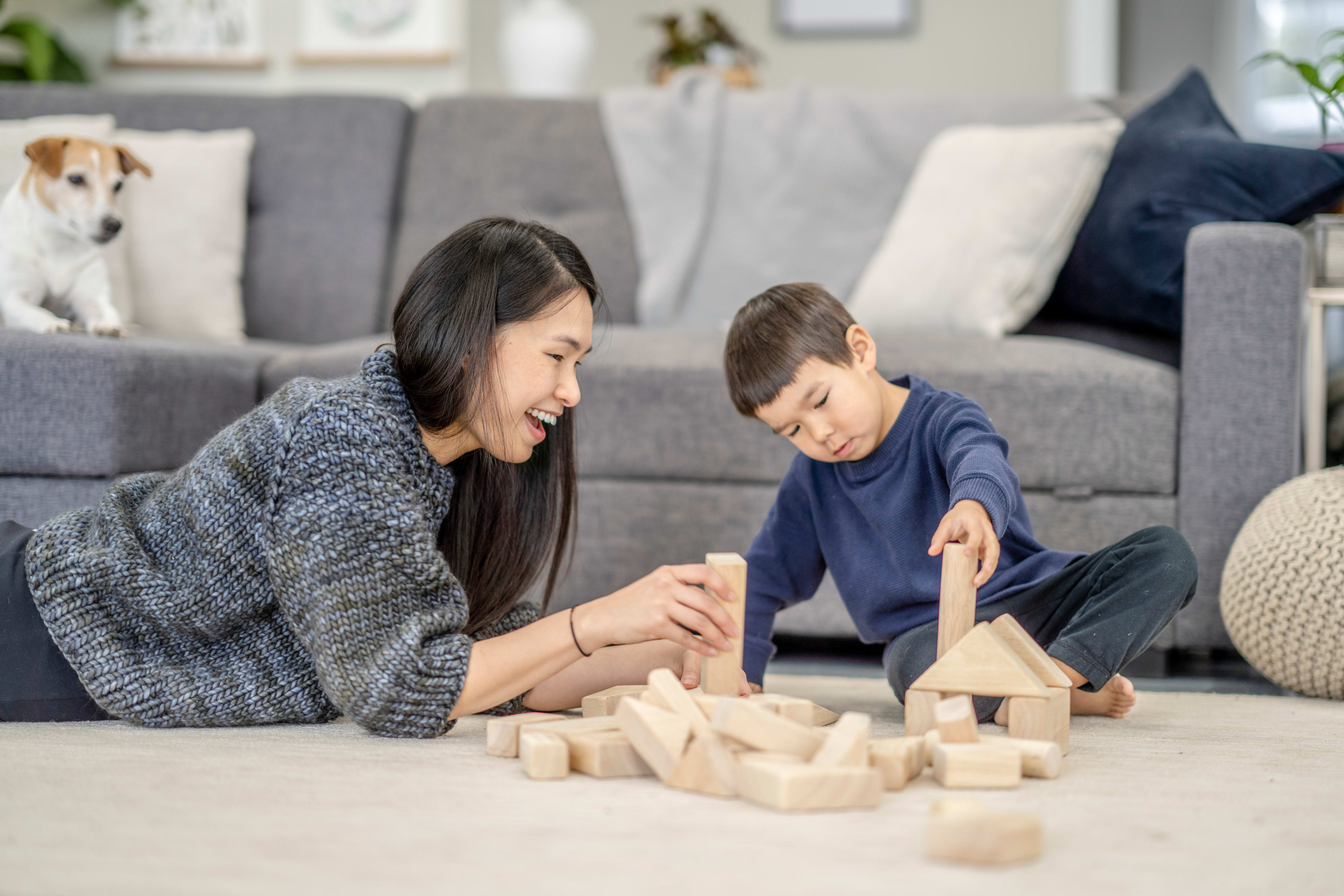 A woman and a young boy are sitting on the floor, playing with wooden blocks in a cozy living room setting with a dog in the background.
