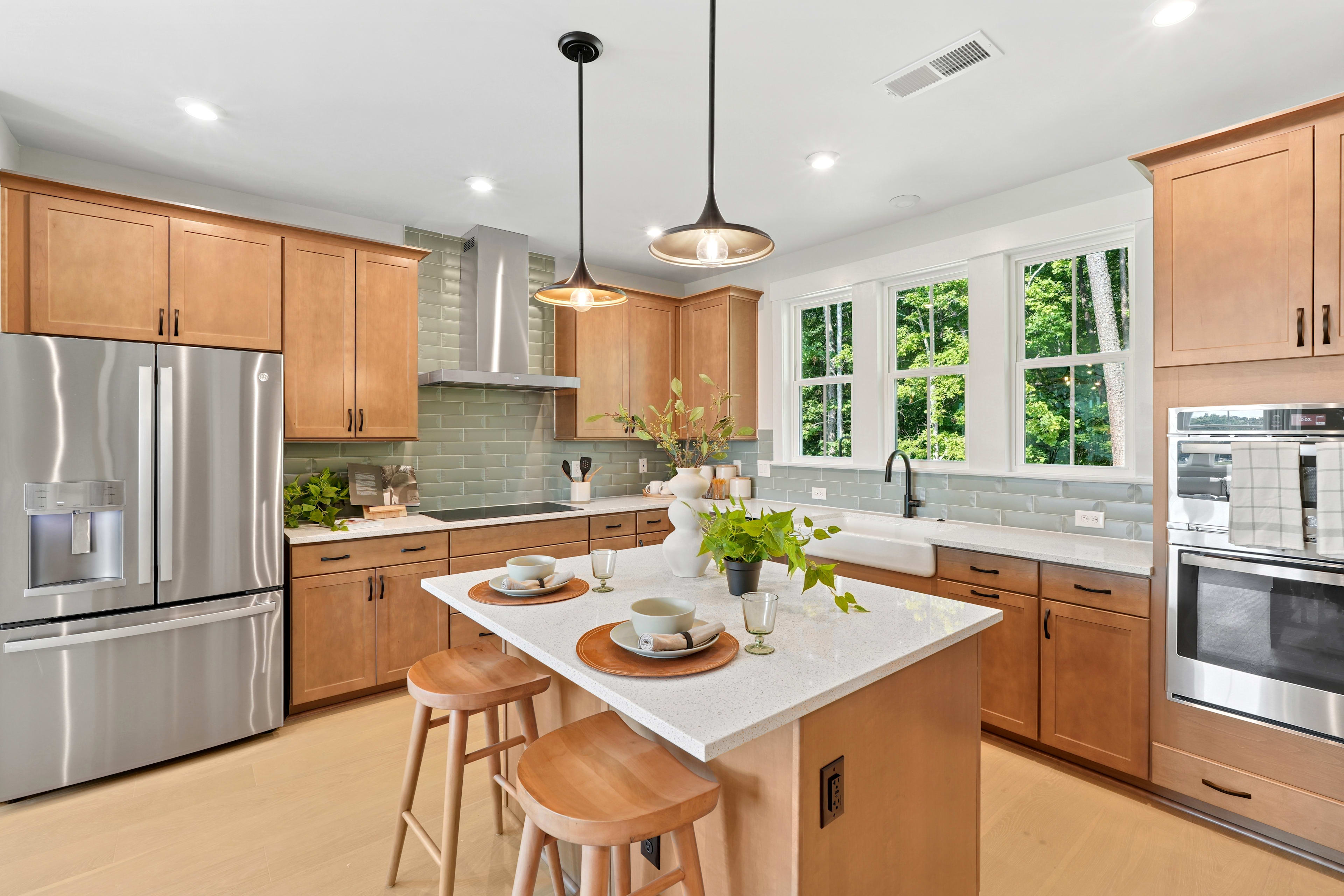 A modern, well-lit kitchen with wooden cabinets, a white countertop, and large windows overlooking a lush outdoor area.