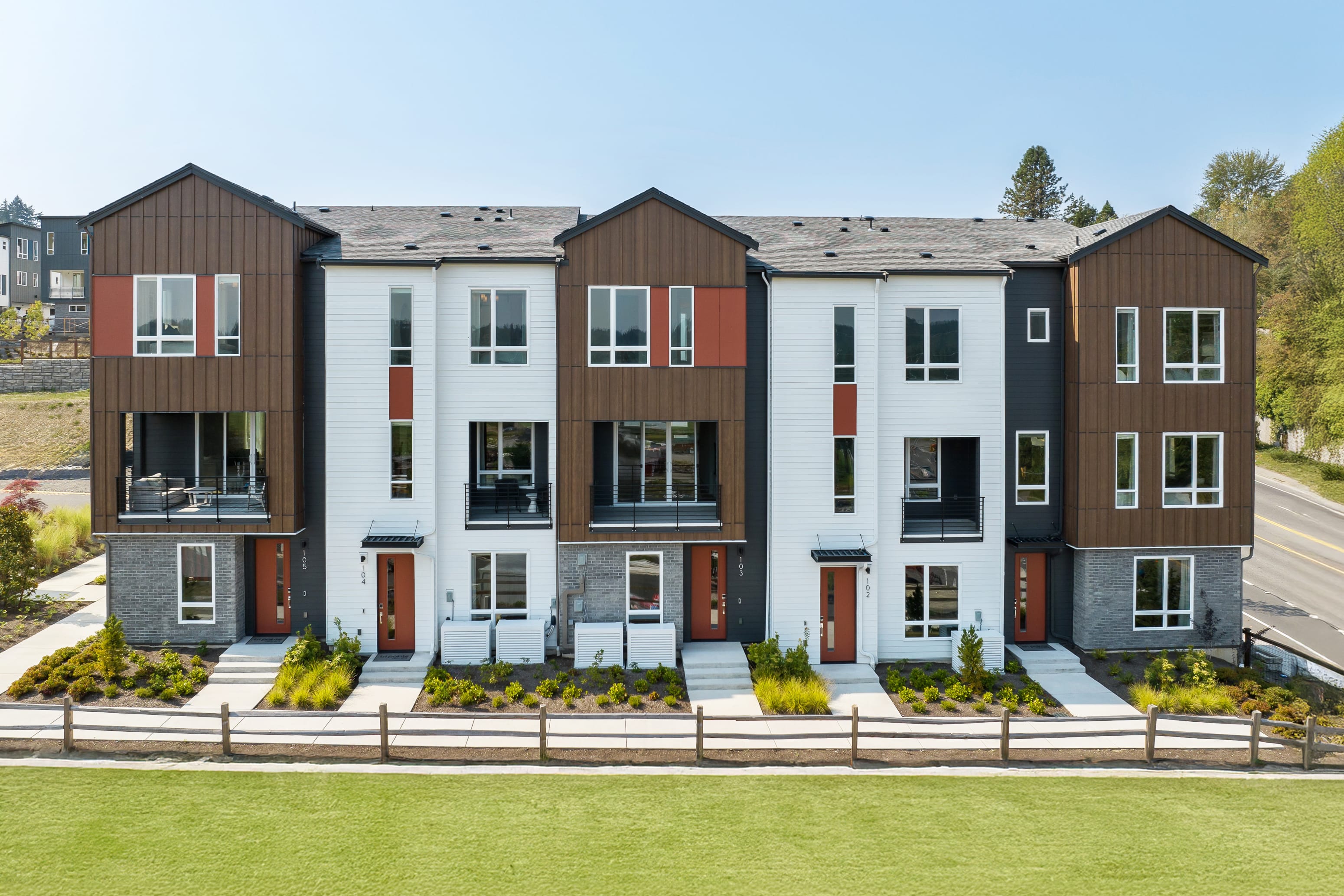 A row of modern, multi-story townhouses with a well-manicured lawn in the foreground, set against a backdrop of trees and a clear blue sky.