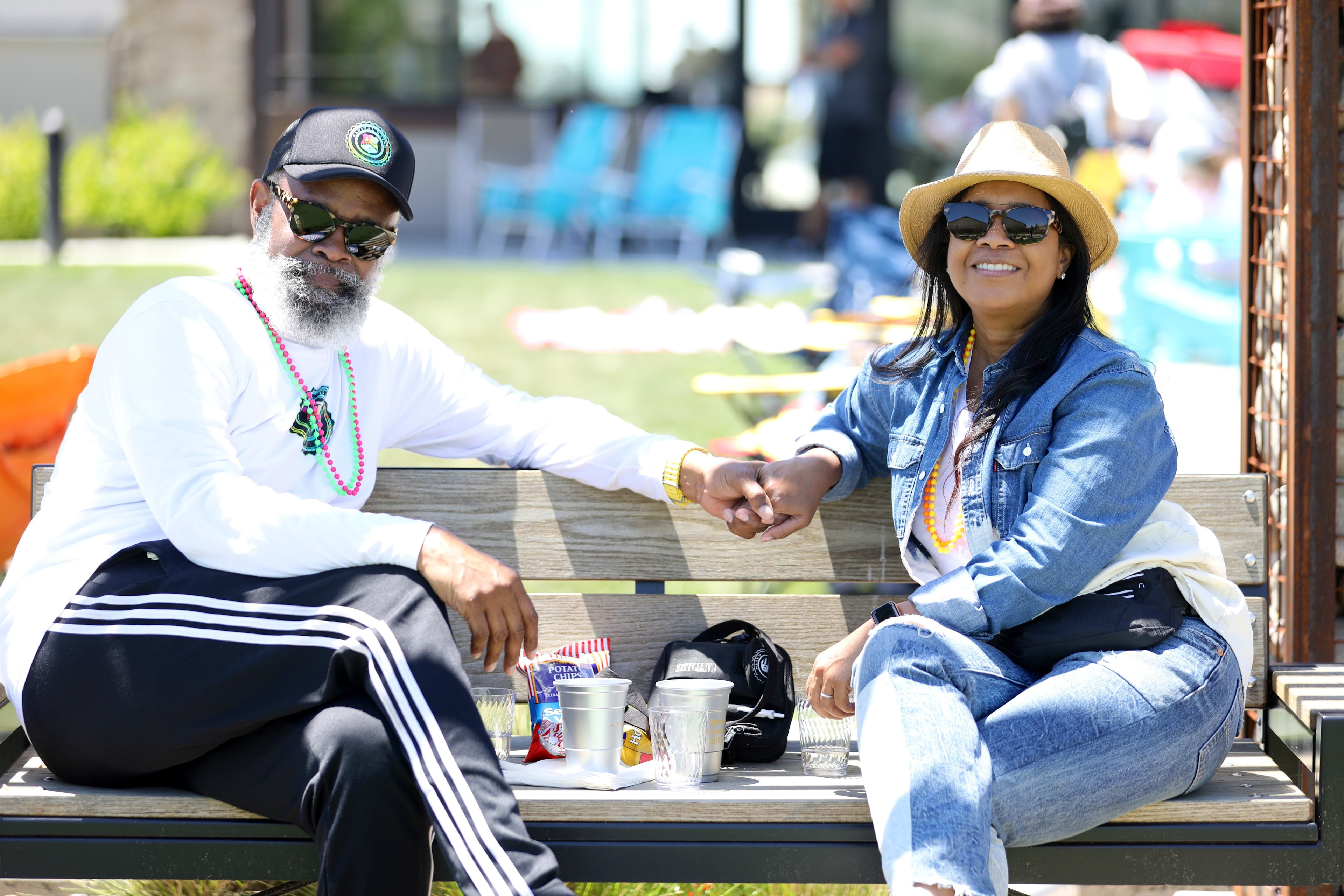 Two people, a man with a long beard and a woman wearing sunglasses, are sitting on a bench in an outdoor setting with people walking by in the background.