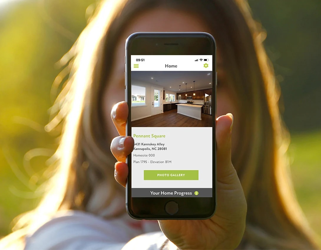 A person's hand is holding a smartphone displaying an interior design app, with a blurred natural background of greenery and sunlight.