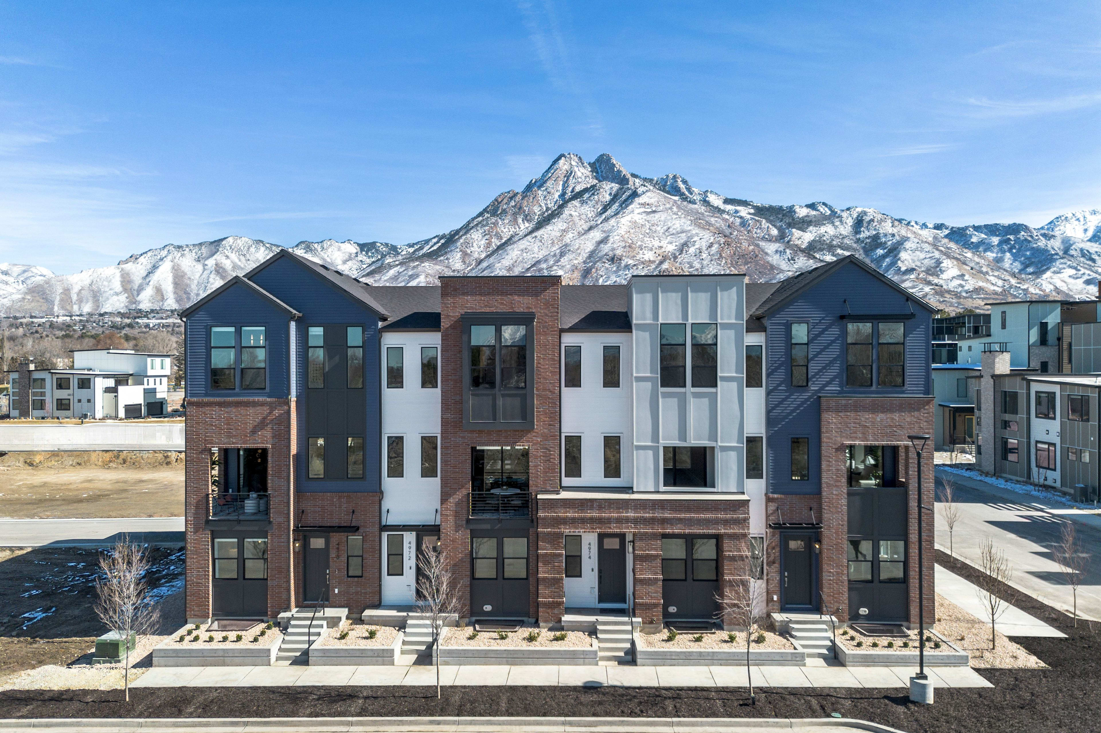 A modern multi-story residential building with brick and glass facades stands in the foreground, with majestic snow-capped mountains visible in the background under a clear blue sky.