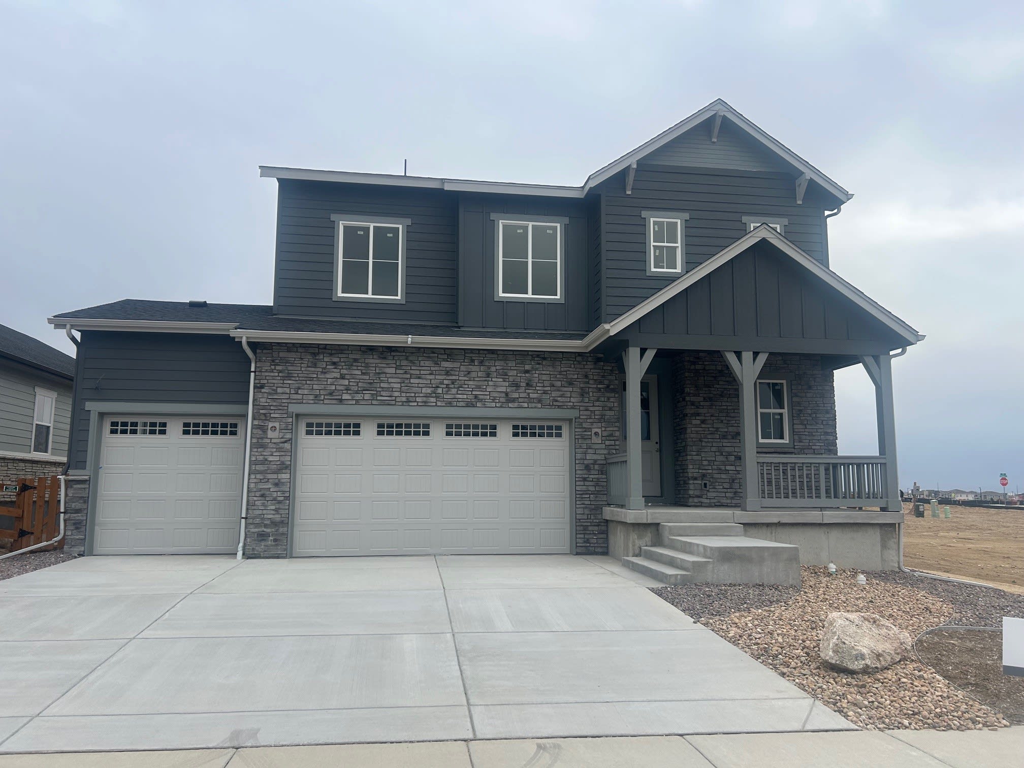 A two-story house with a gray exterior, stone accents, and a garage door in the foreground, set against a cloudy sky in the background.