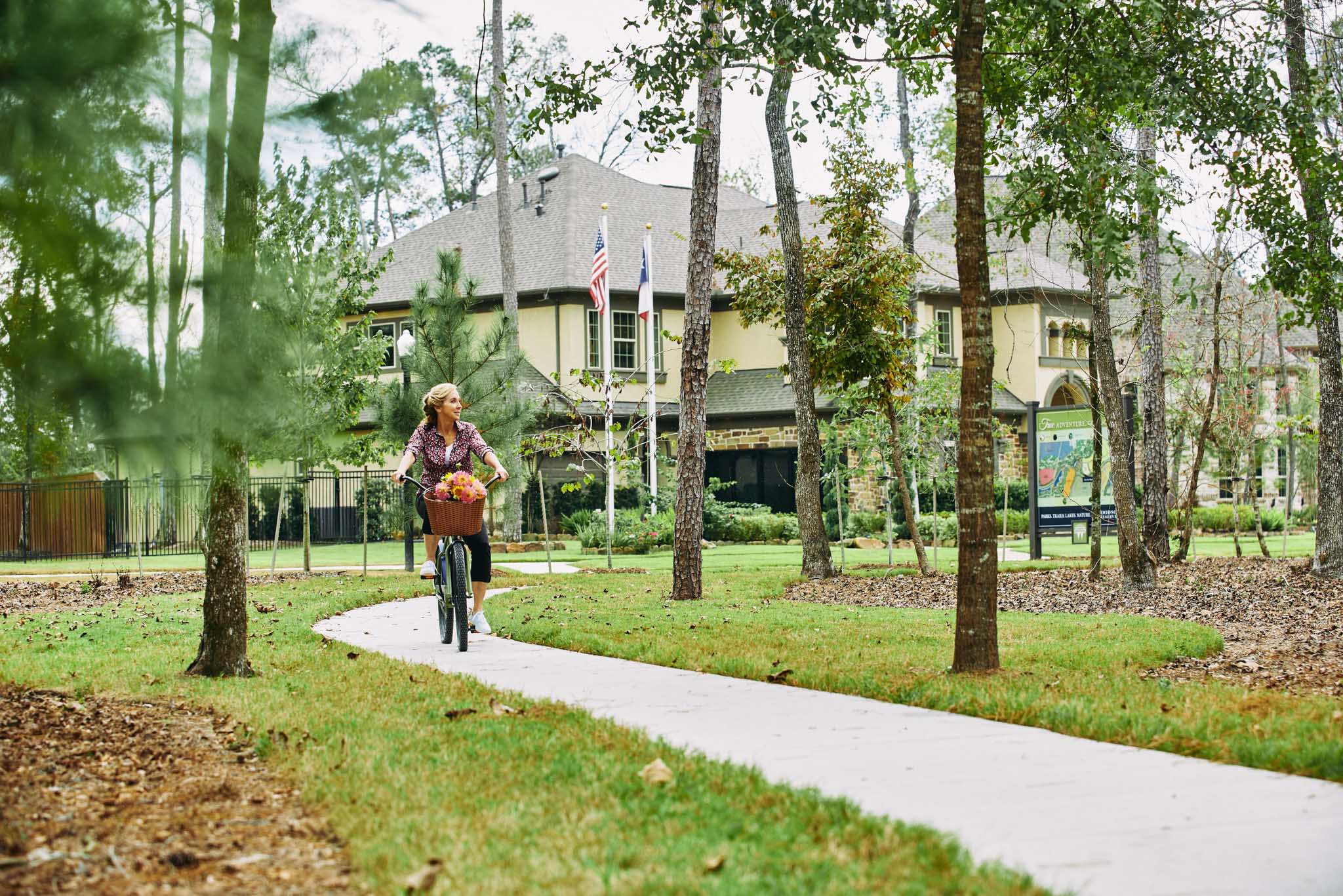 A person is walking along a paved path through a wooded area, with a multi-story building visible in the background.