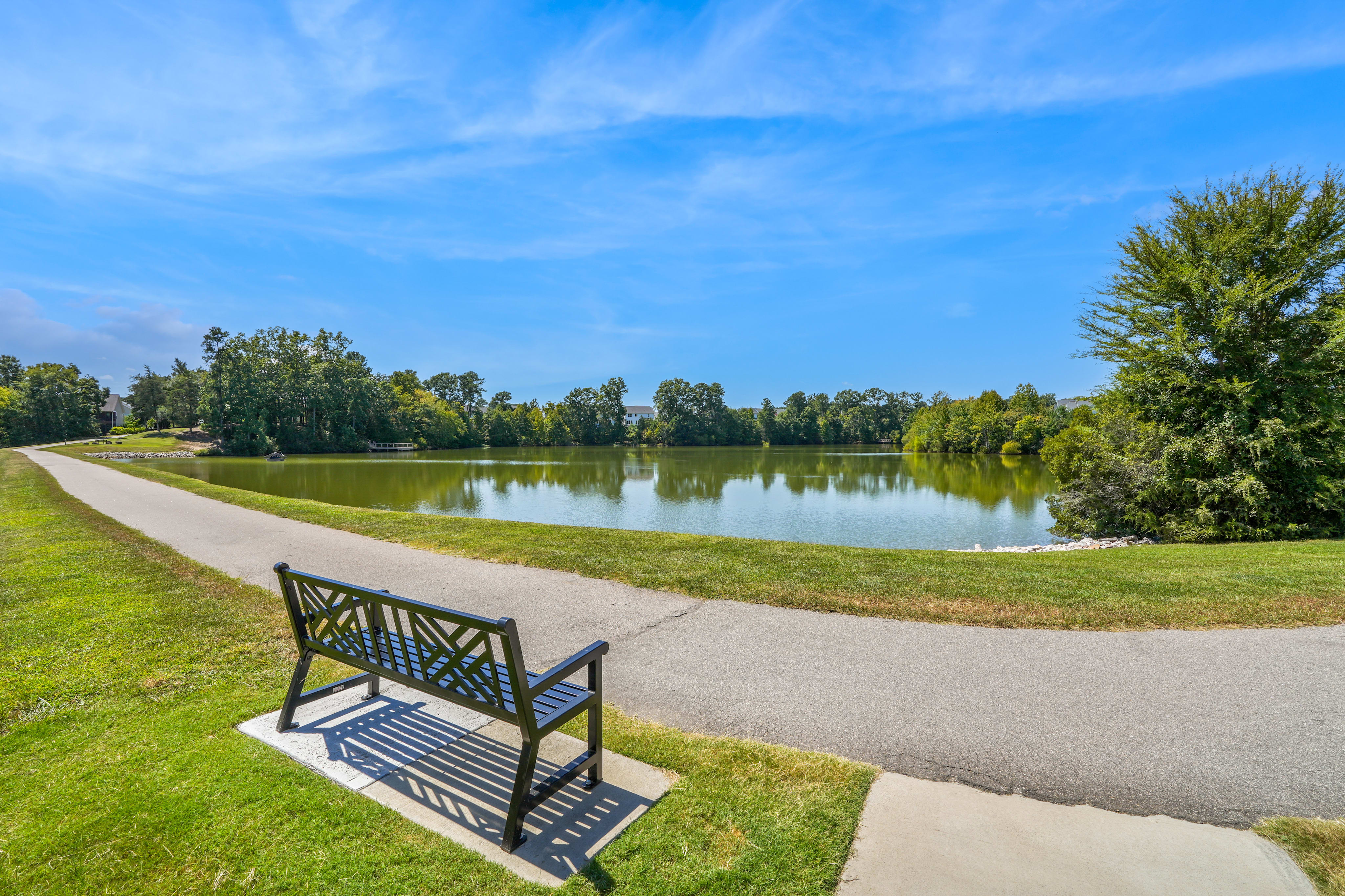 A peaceful park setting with a paved walkway leading along a tranquil lake surrounded by lush greenery and a clear blue sky.
