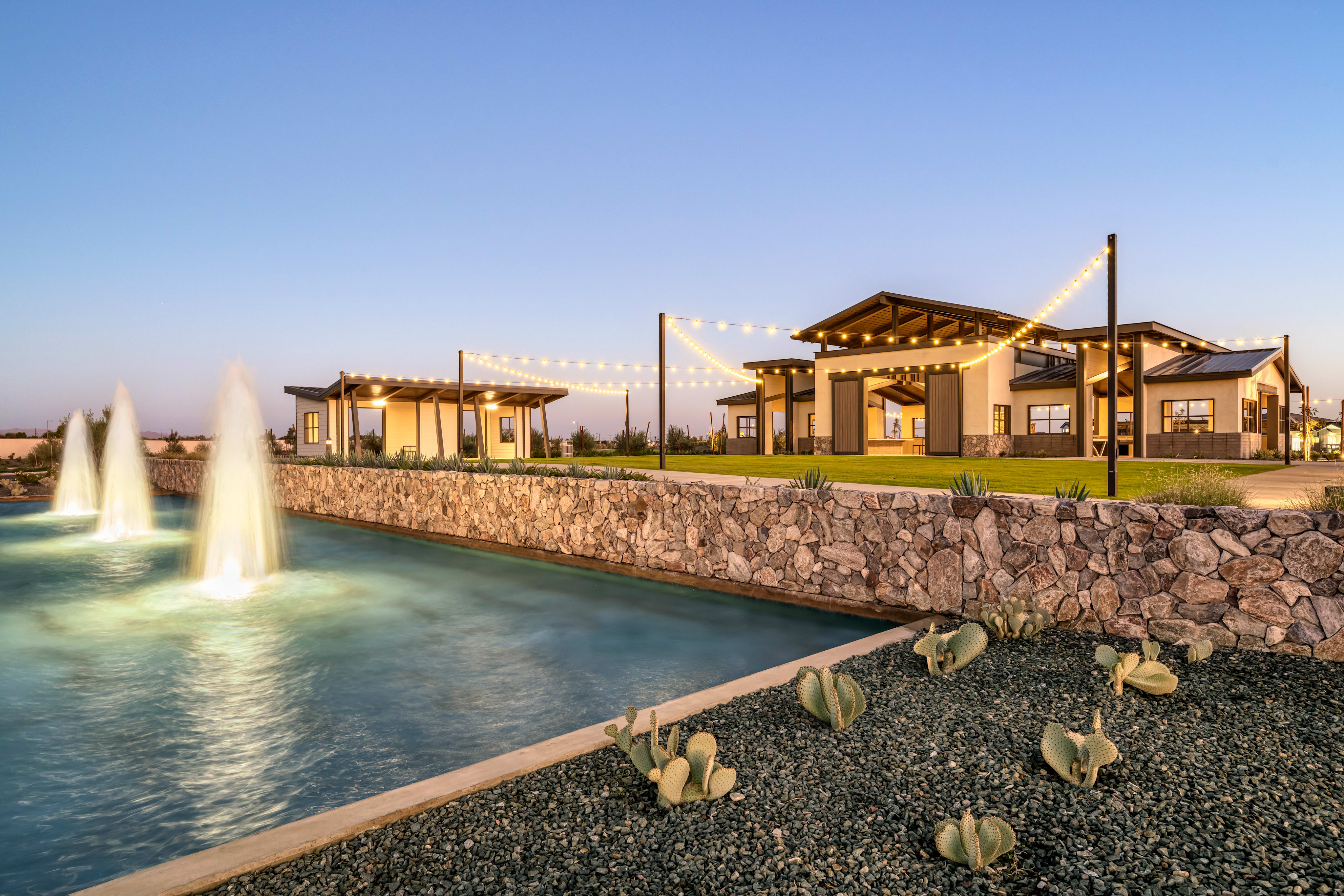 A large, modern building with a stone exterior and wooden accents stands in the background, surrounded by a lush green lawn. In the foreground, a tranquil water feature with fountains cascades over a rocky landscape, creating a serene and picturesque scene.