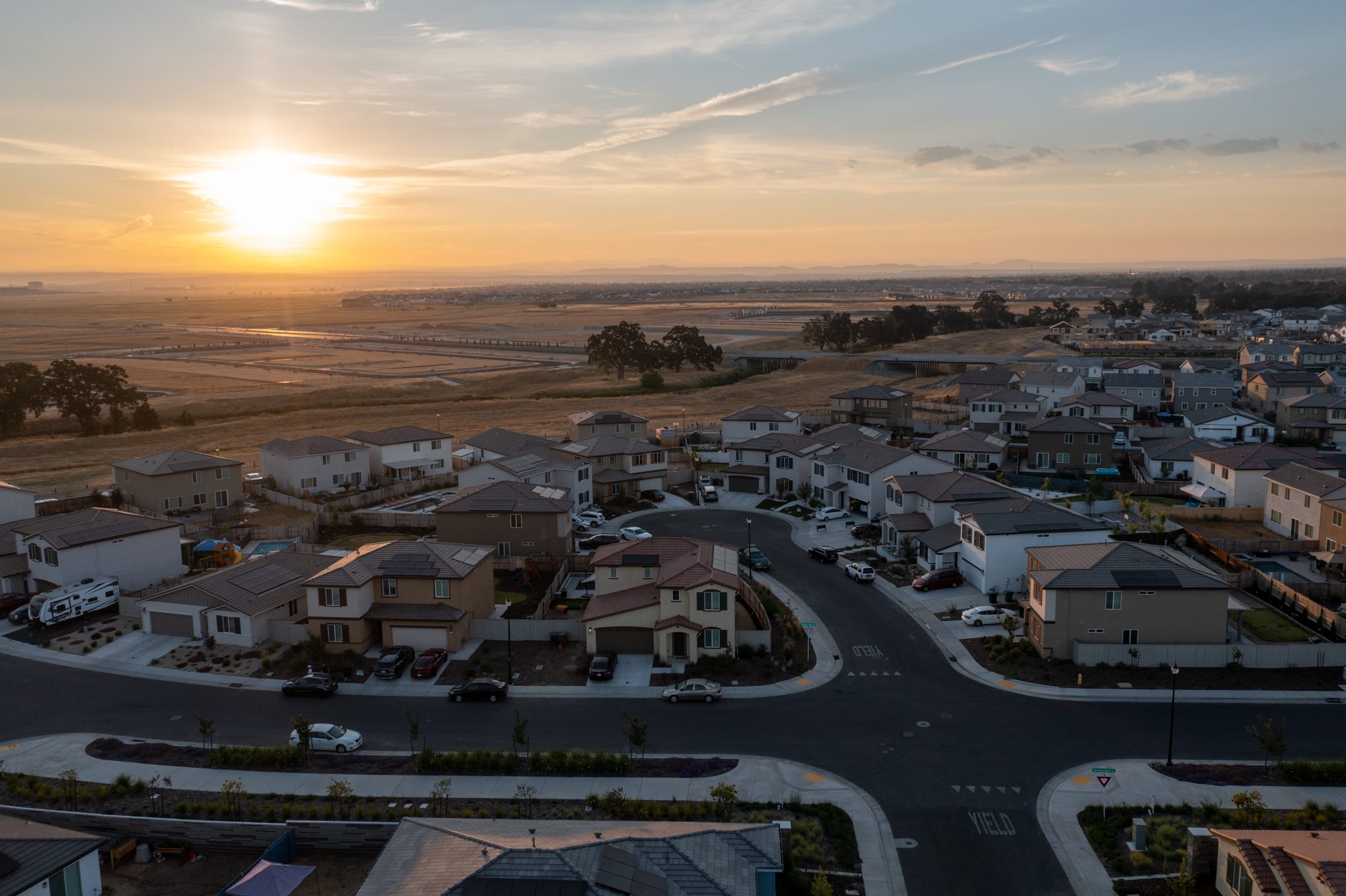 A picturesque suburban neighborhood nestled amidst rolling hills, with a stunning sunset illuminating the sky and casting a warm glow over the rooftops and streets below.