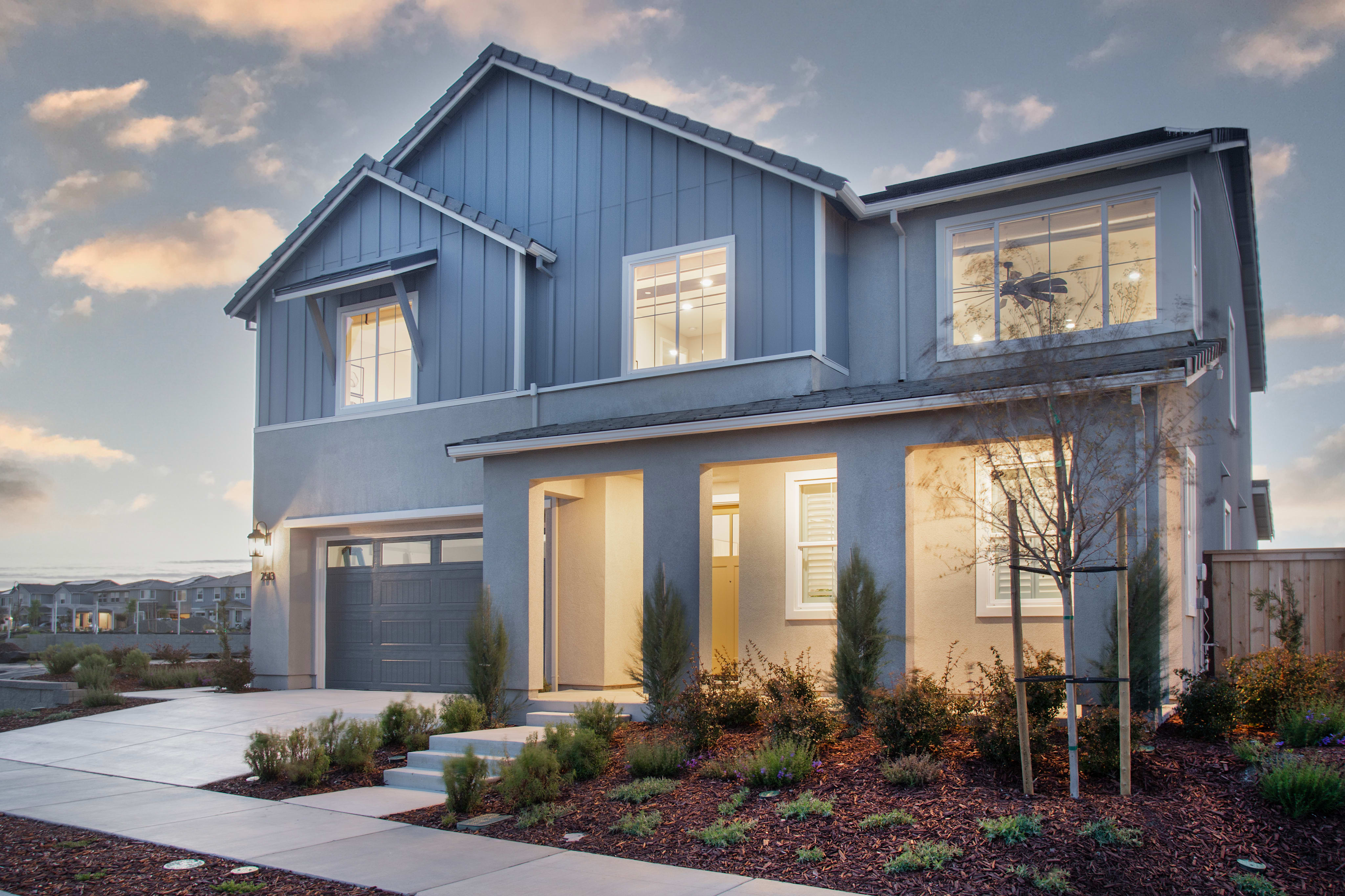 A modern two-story house with a gray exterior, a garage door, and a well-landscaped front yard set against a cloudy sky in the background.
