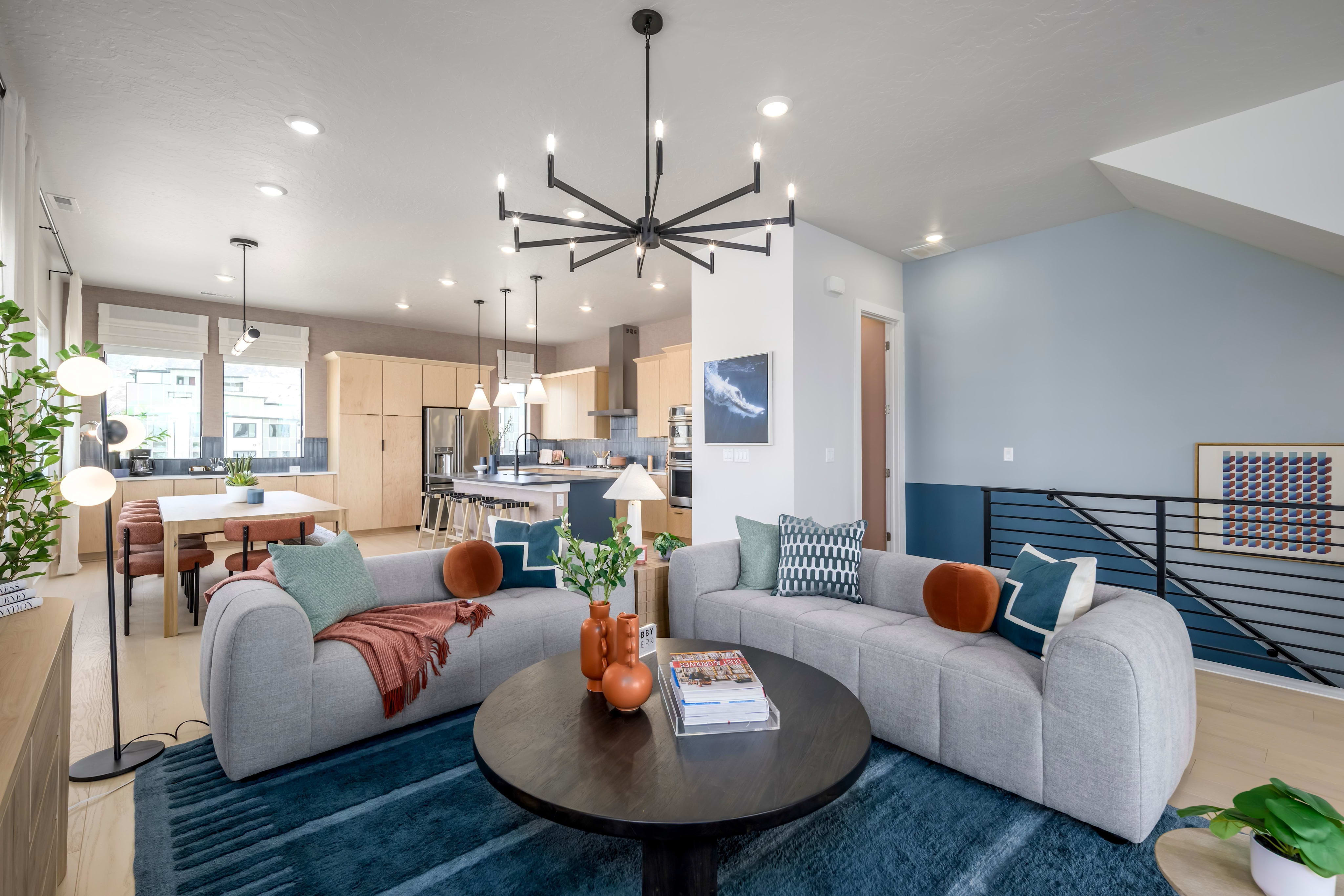 A modern and stylish living room with a large circular coffee table, plush gray sofas, and a striking black chandelier overhead, complemented by a kitchen area visible in the background.