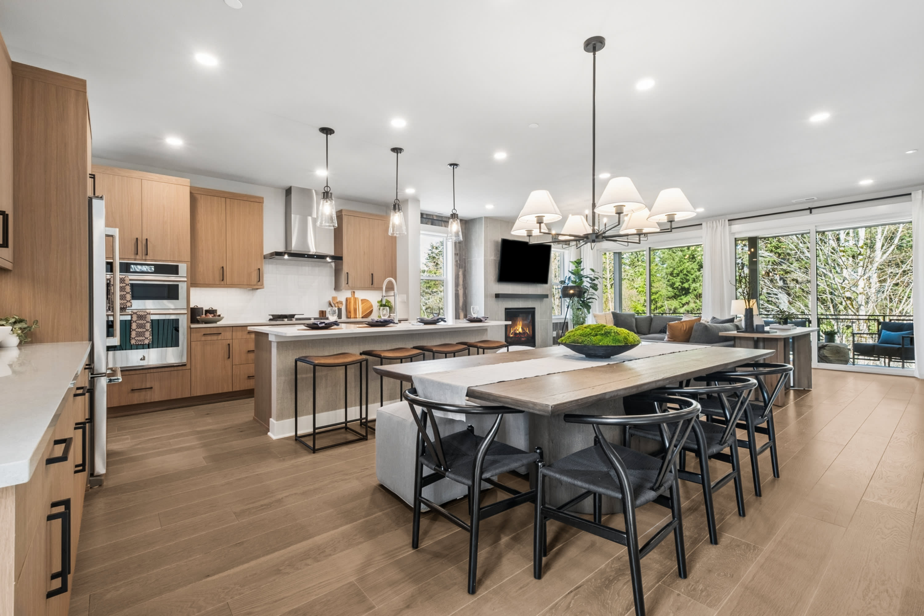 A modern and spacious kitchen with a large island, wooden cabinets, and pendant lighting fixtures, set against a backdrop of a cozy living area with a TV and large windows overlooking the outdoors.