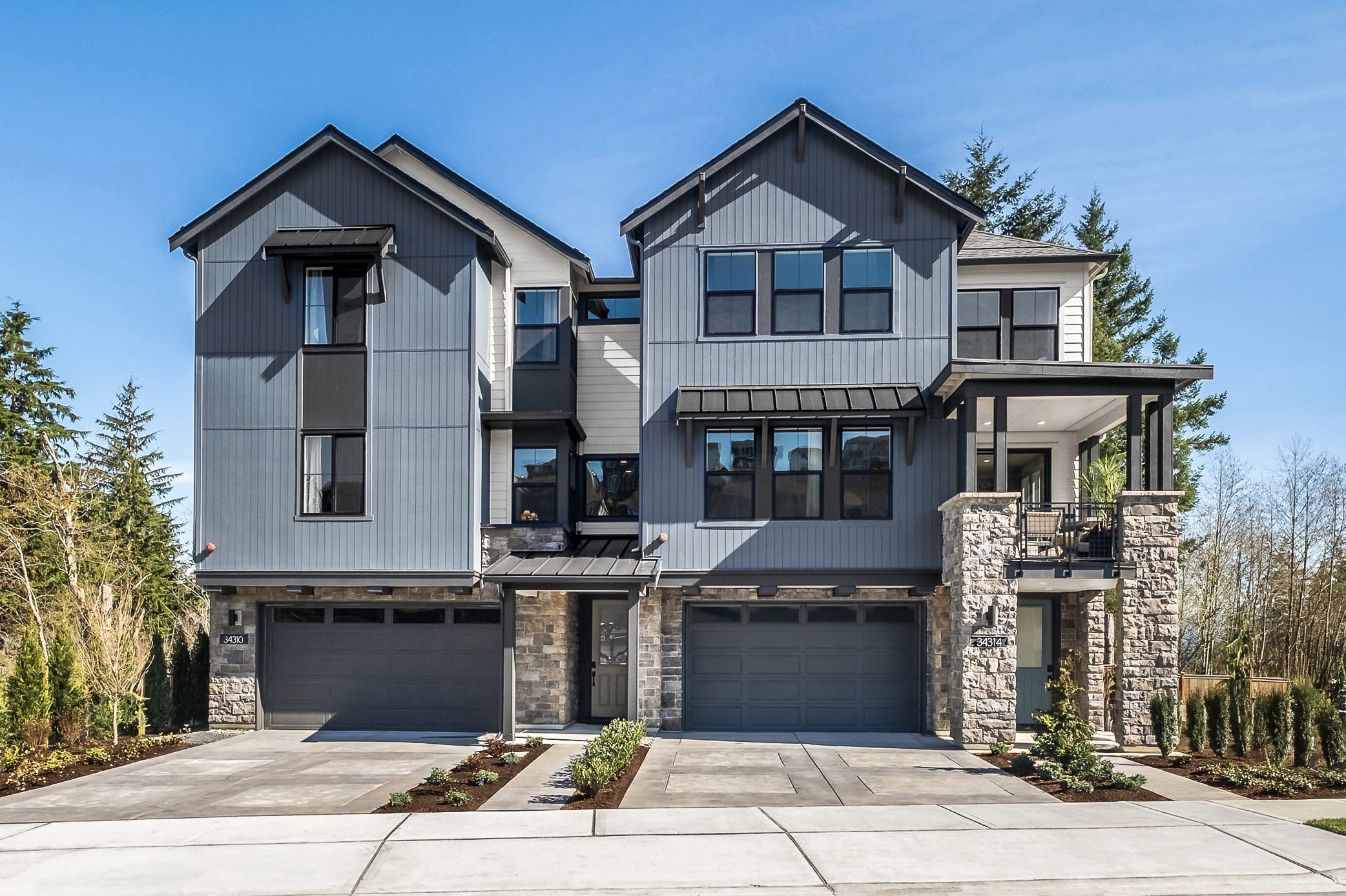 The image depicts a modern, multi-story residential building with a gray exterior, large windows, and a stone foundation. The building is surrounded by trees and greenery, suggesting a suburban or rural setting.