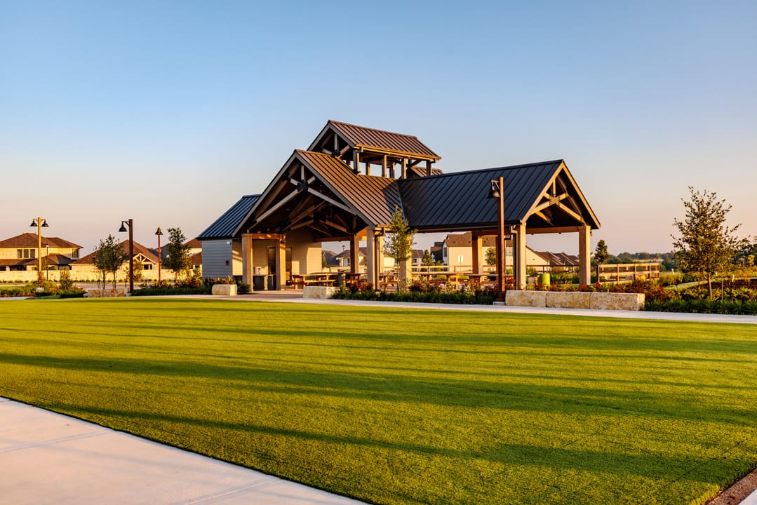 A vibrant green lawn stretches towards a modern pavilion with a dark metal roof, set against a backdrop of houses and trees under a clear sky.