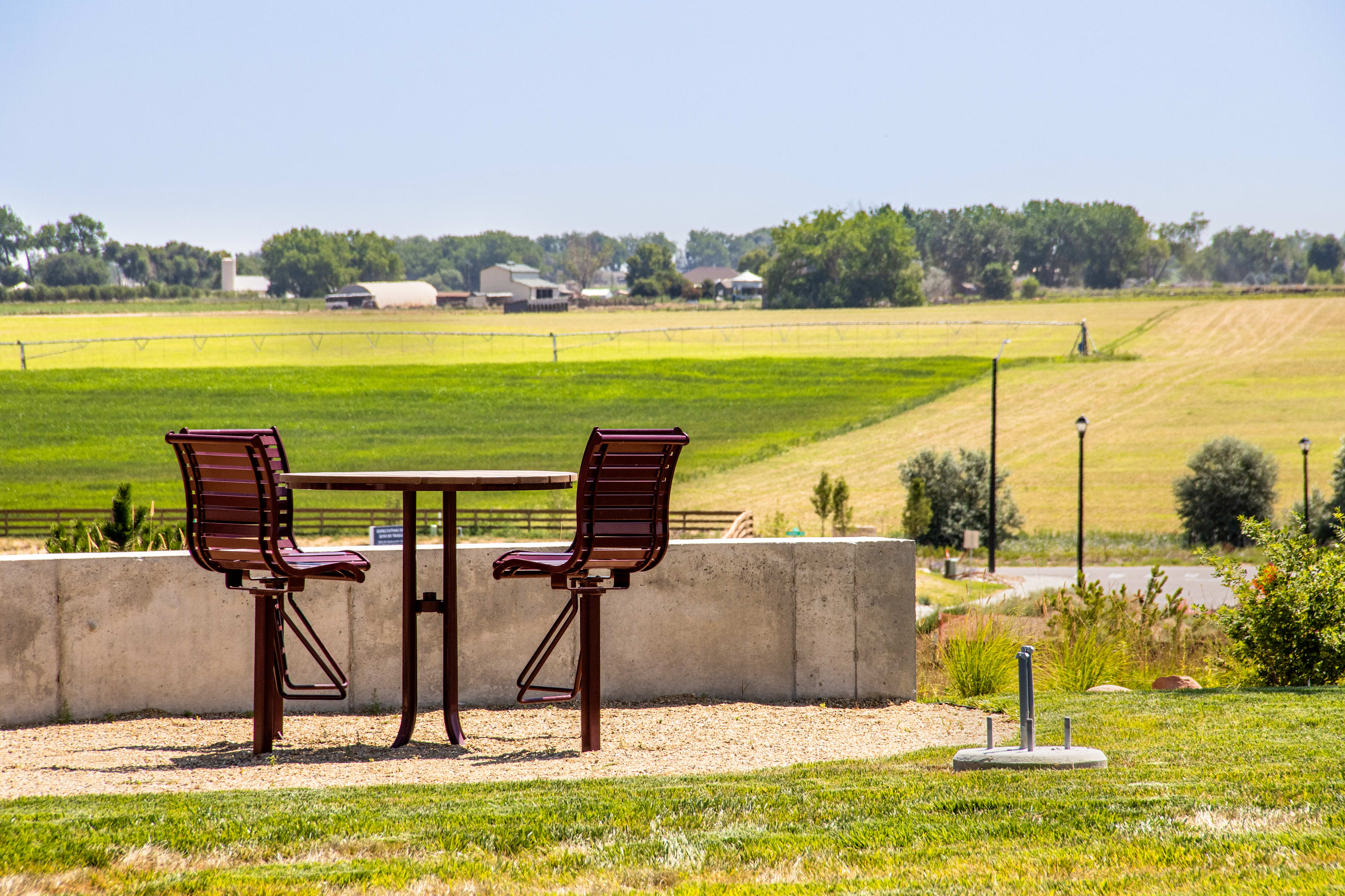 Two red bar stools surround a small table on a gravel patio in front of a concrete wall, with a vast, green, and yellow rural landscape stretching into the distance under a clear blue sky.