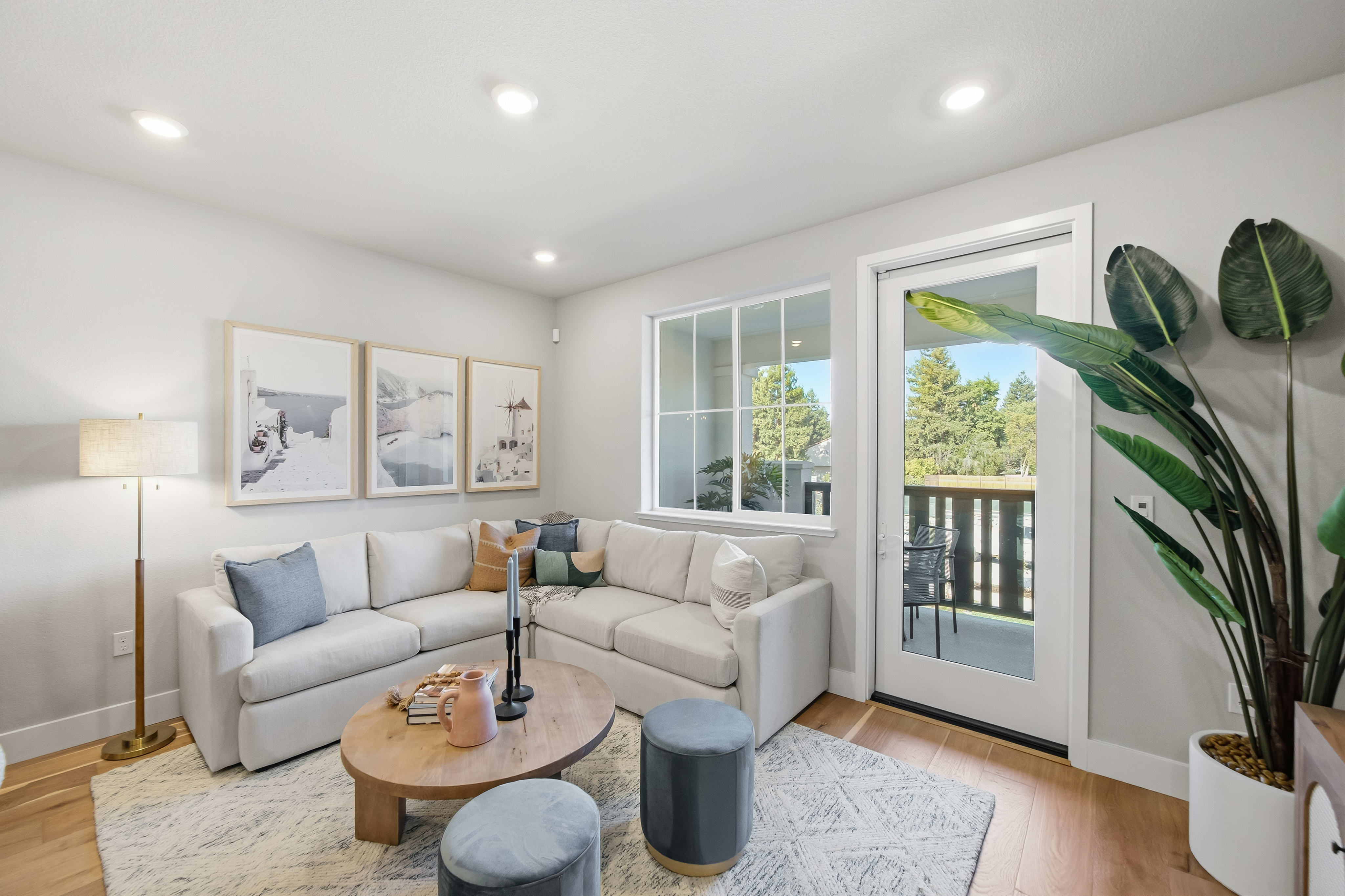 A modern living room with a sectional sofa and coffee table in the foreground, and a glass door leading to an outdoor patio with trees visible in the background.
