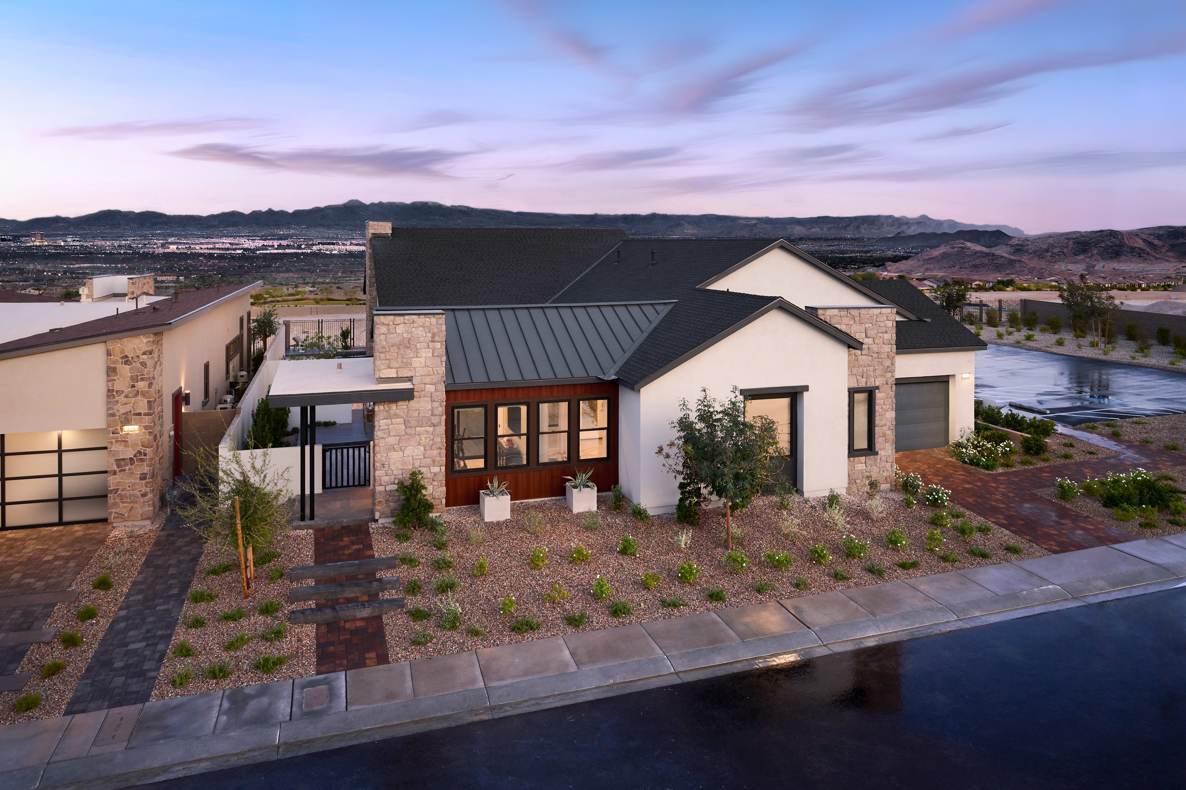 A modern, multi-story residential building with a stone and metal exterior sits amidst a landscaped garden, overlooking a body of water and distant mountains in the background.
