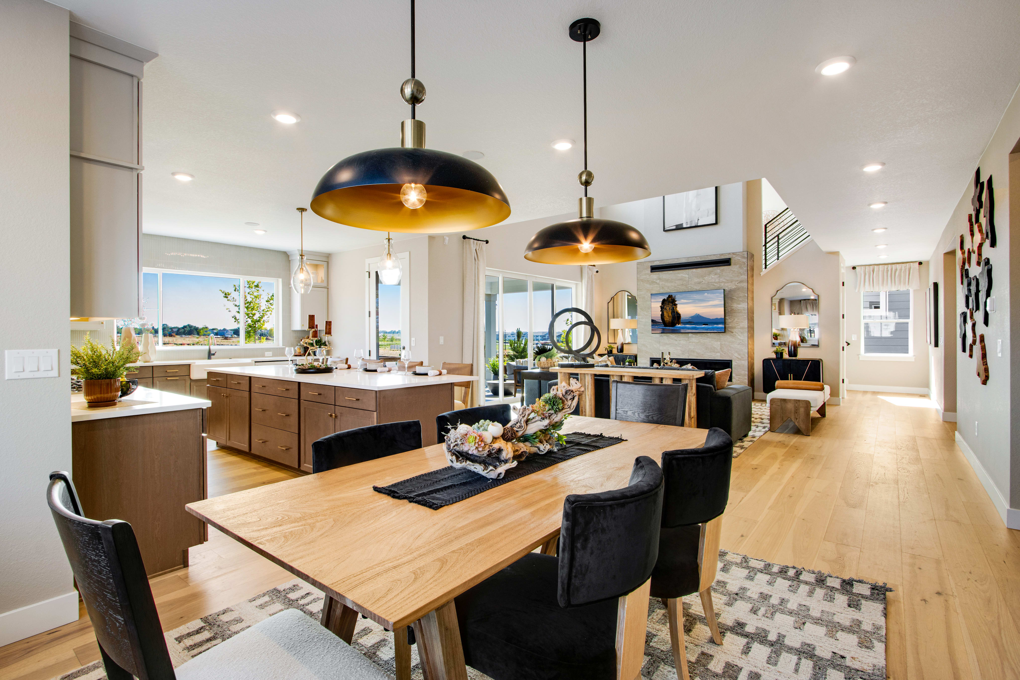 A modern dining room with a wooden table and chairs in the foreground opens to a spacious living area with a fireplace and large windows in the background.