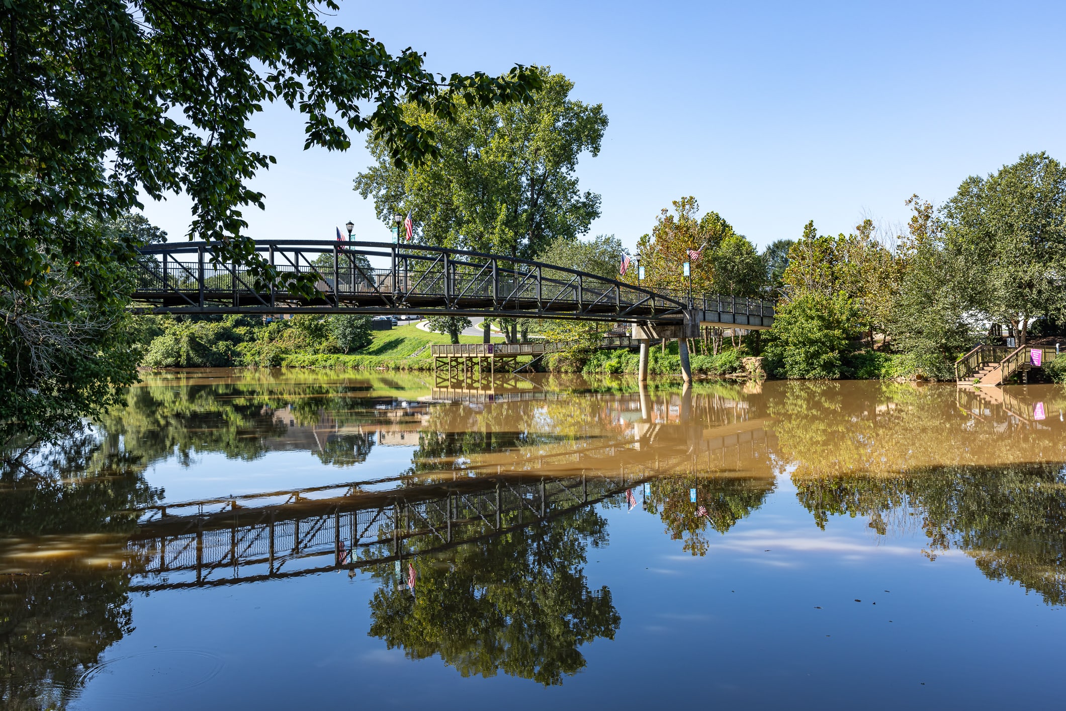 A metal footbridge spans a calm river reflecting the lush green trees and blue sky in the background.