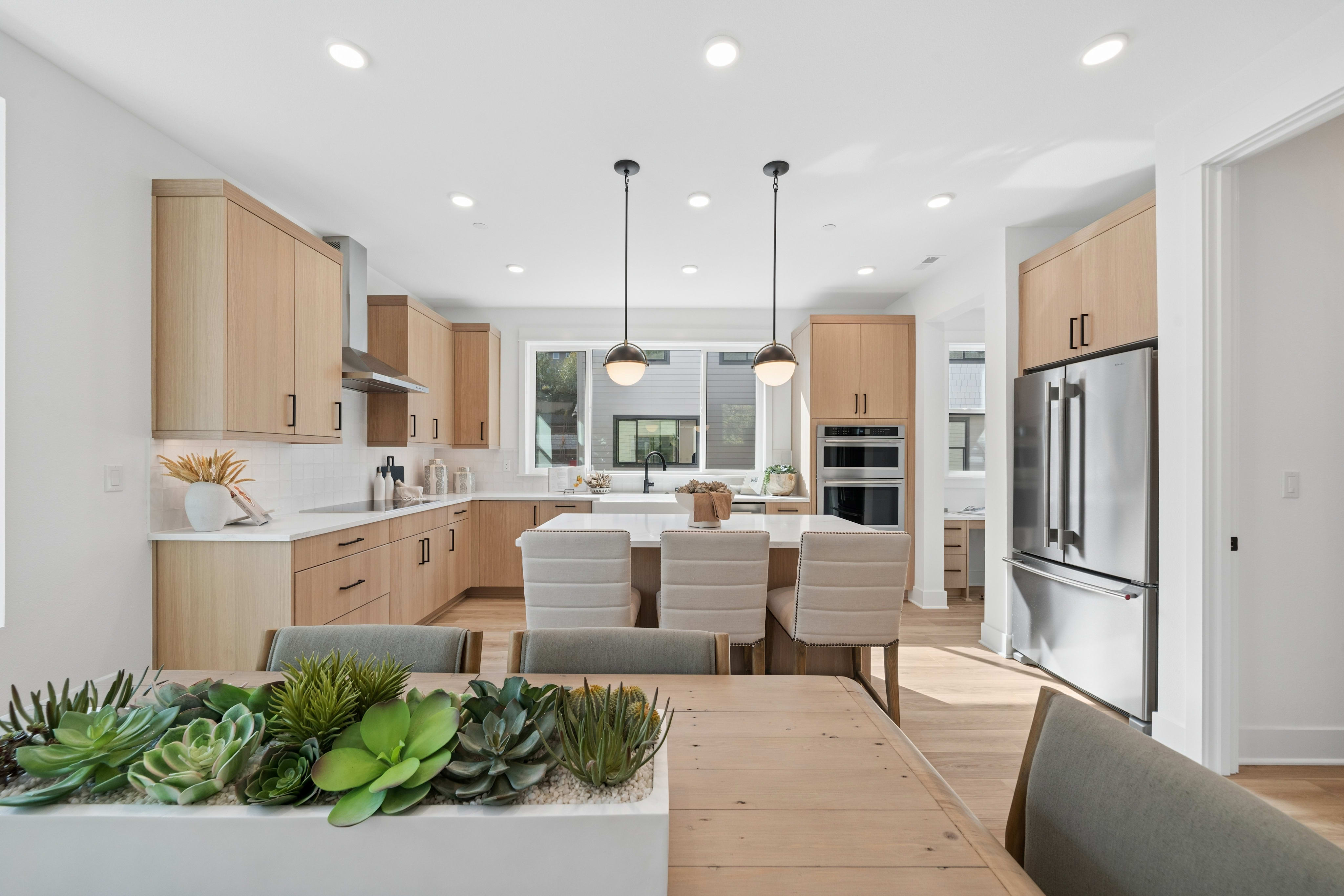 A wooden table with a succulent planter sits in the foreground of a modern kitchen featuring light wood cabinetry, a white island with seating, and stainless steel appliances.