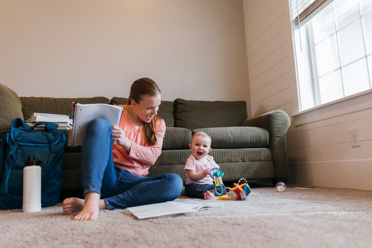 Infant and mother play with toys on living room floor while mother studies