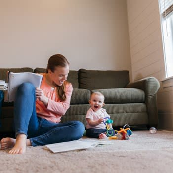 Infant and mother play with toys on living room floor while mother studies