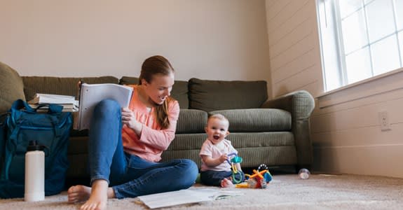 Infant and mother play with toys on living room floor while mother studies